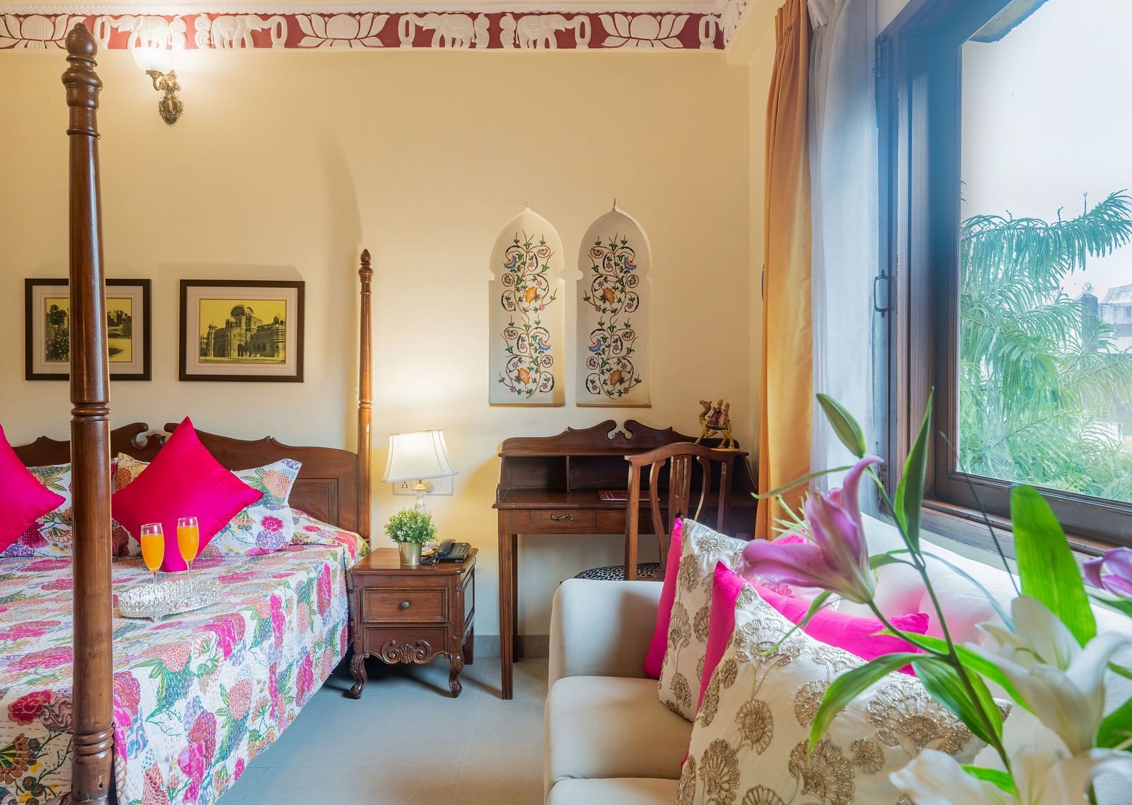 A lovely room at Dera Rawatsar, Jaipur, features a four-poster bed, a comfortable sofa, a writing desk, and decorative floral accents on the wall.
