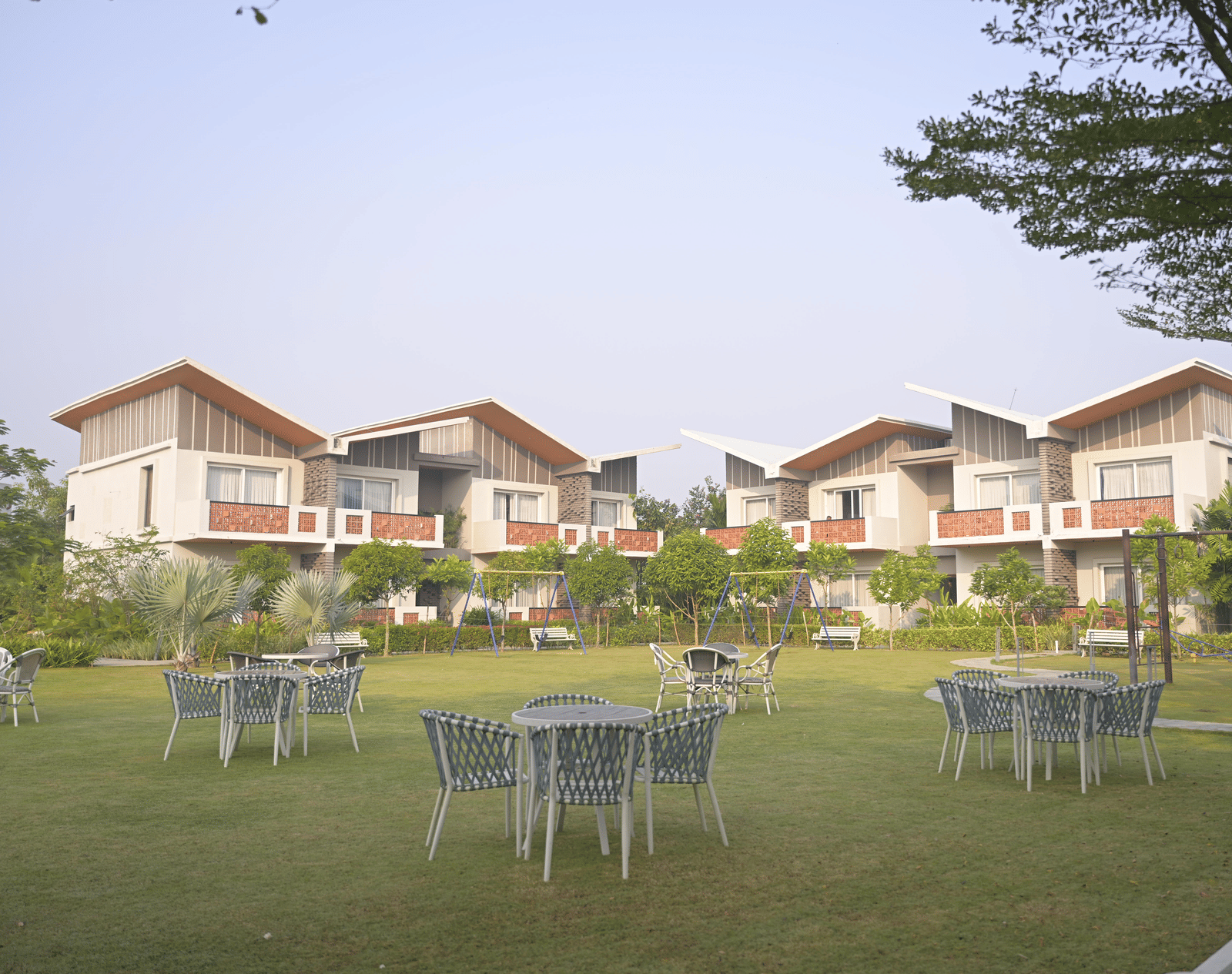 Open green lawn with white outdoor seating and rows of villas in the distance at Hotel Sonar Bangla Mayapur.