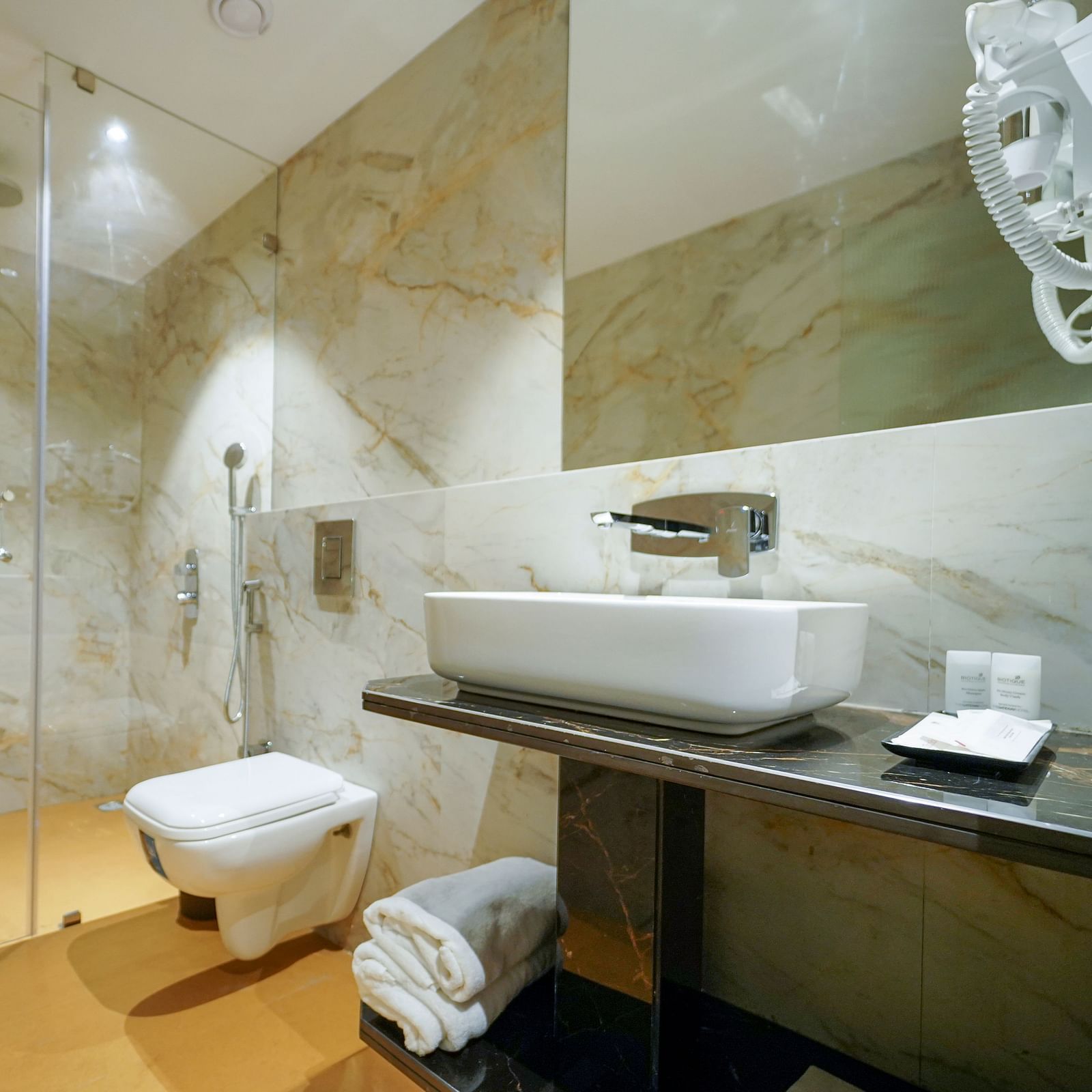 A bathroom features a glass-enclosed shower area, a toilet, and a sink with a faucet and toiletries on a countertop - Hotel Park Inn, Nellore.