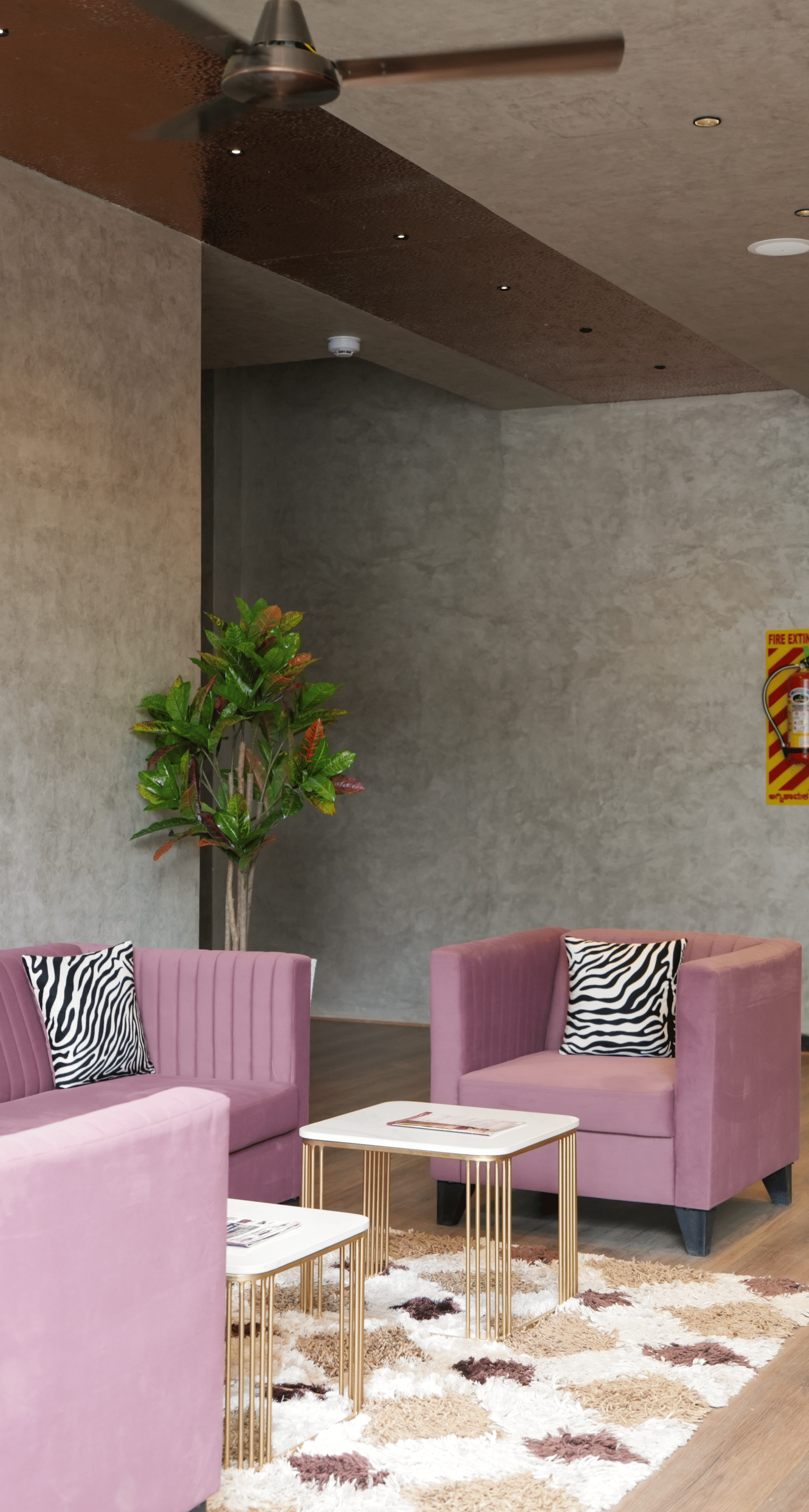 Lounge area with pink sofas, a TV displaying the hotel logo, and decorative plants - TGI Livancia Hillside, JC Nagar, Mysore