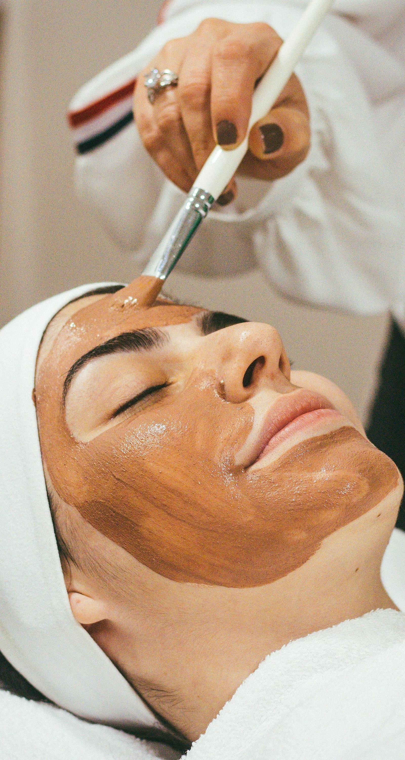 a woman with her eyes closed getting a brown face mask applied on her face