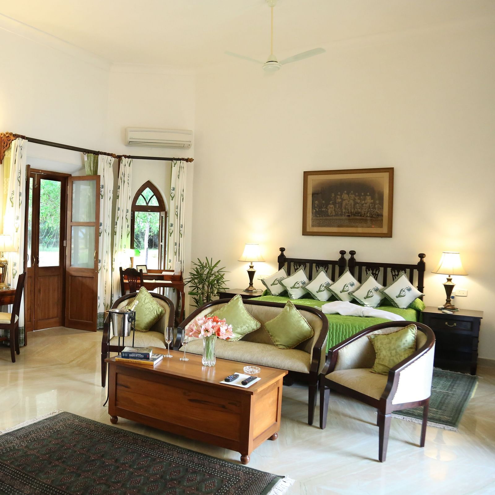 A well-lit room featuring a seating area with a sofa and chairs, a bed with decorative pillows, and multiple doors leading to an outdoor space- Shahpura Bagh.
