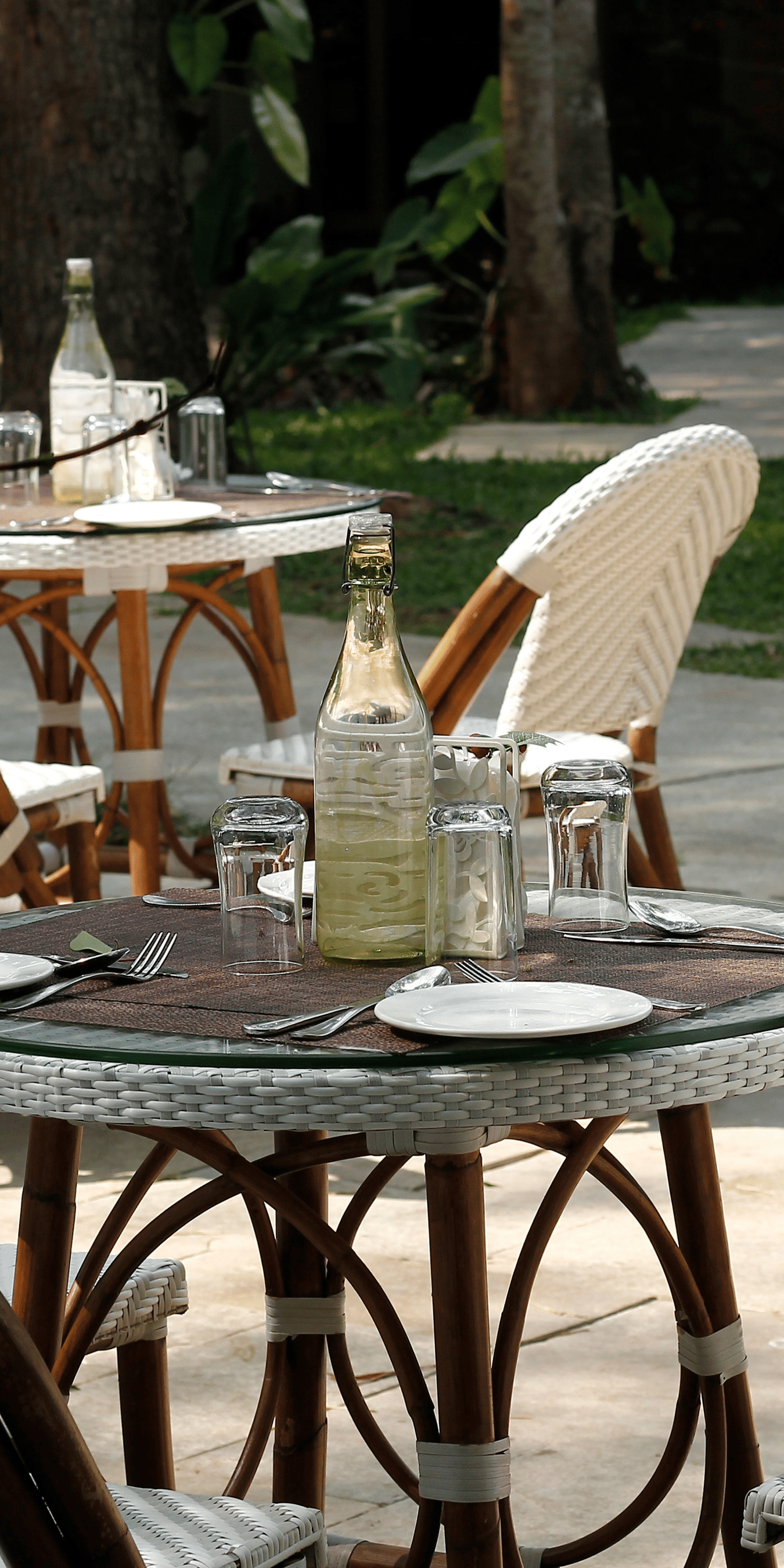 Outdoor cafe setting with wine glasses and a bottle on a table, foliage in the foreground.