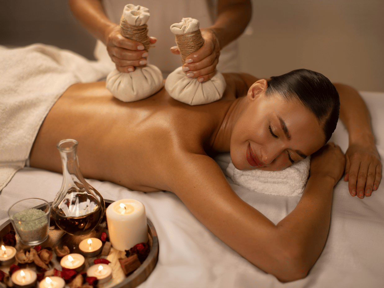 Woman receiving a herbal compress back massage surrounded by spa candles and oils.