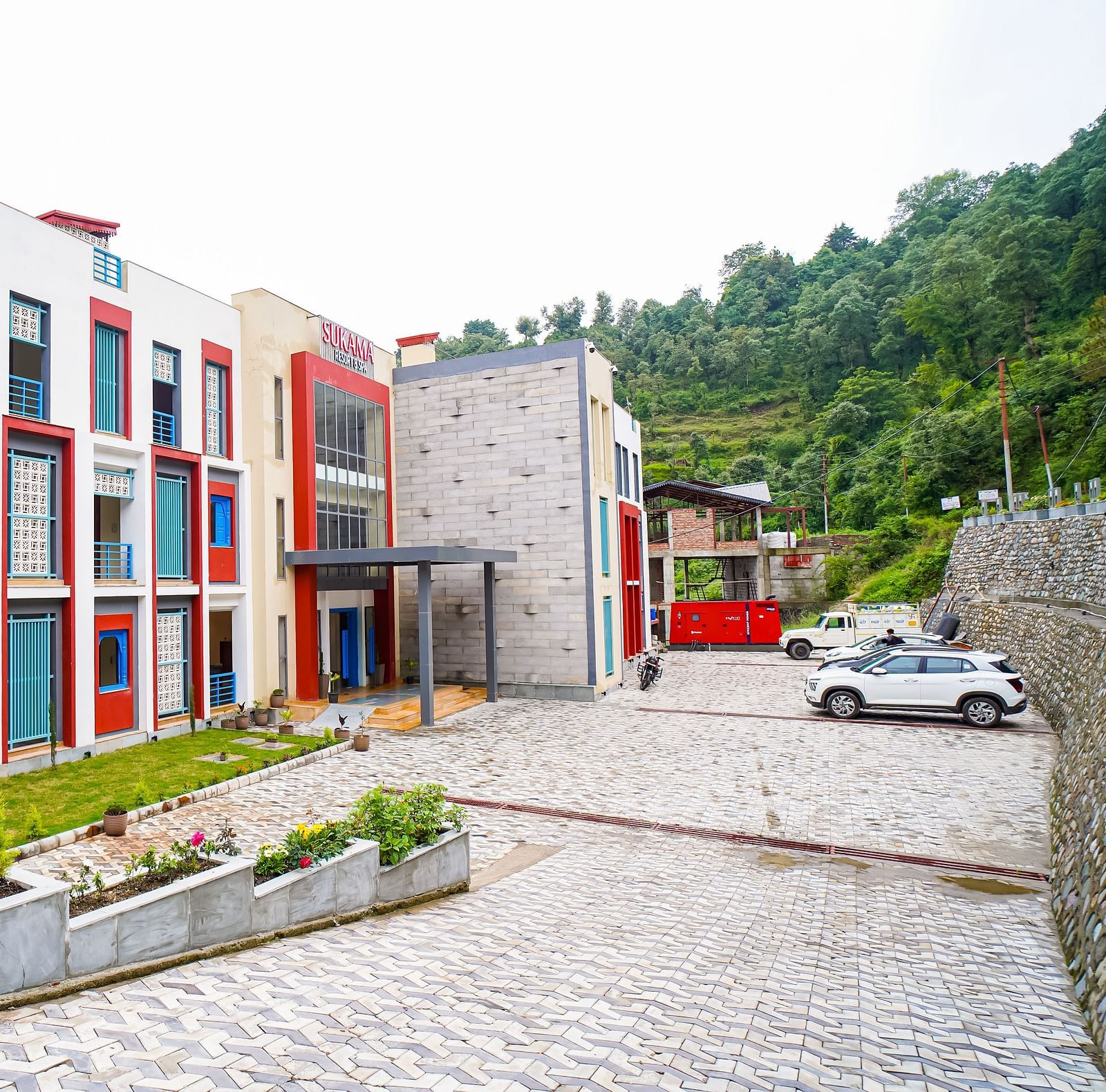 The exterior of a modern building with red and white sections and a paved courtyard is visible | Sukama Resort & Spa, Nainital