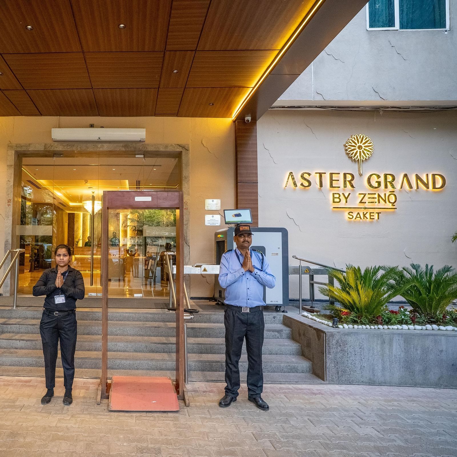 Entrance of ASTER GRAND BY ZENQ SAKET with a car, plants and two staff members standing in the front by joining their hands.