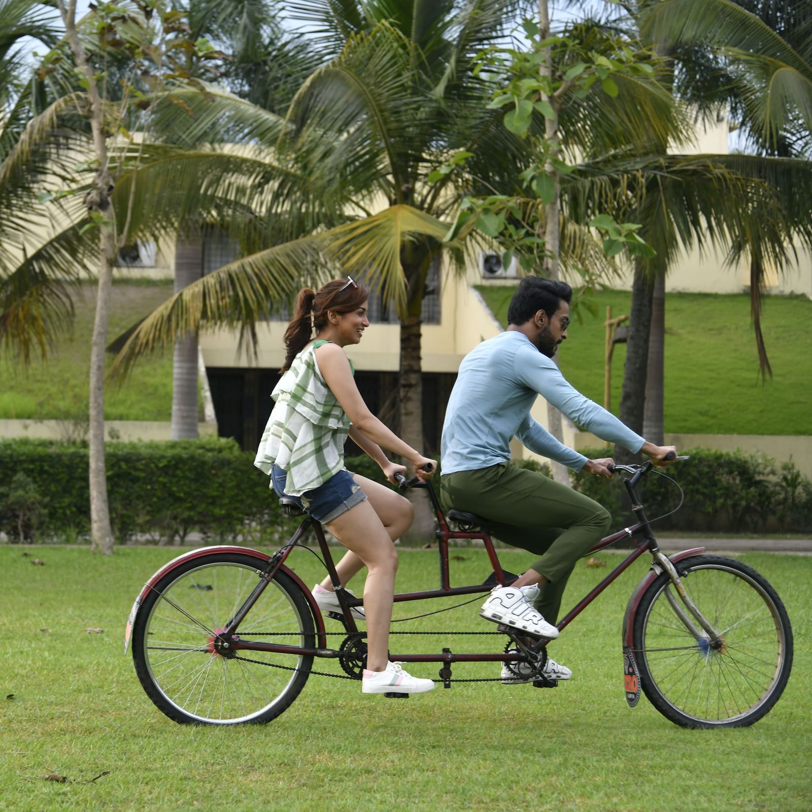 A couple rides a tandem bicycle on a grassy lawn with palm trees in the background