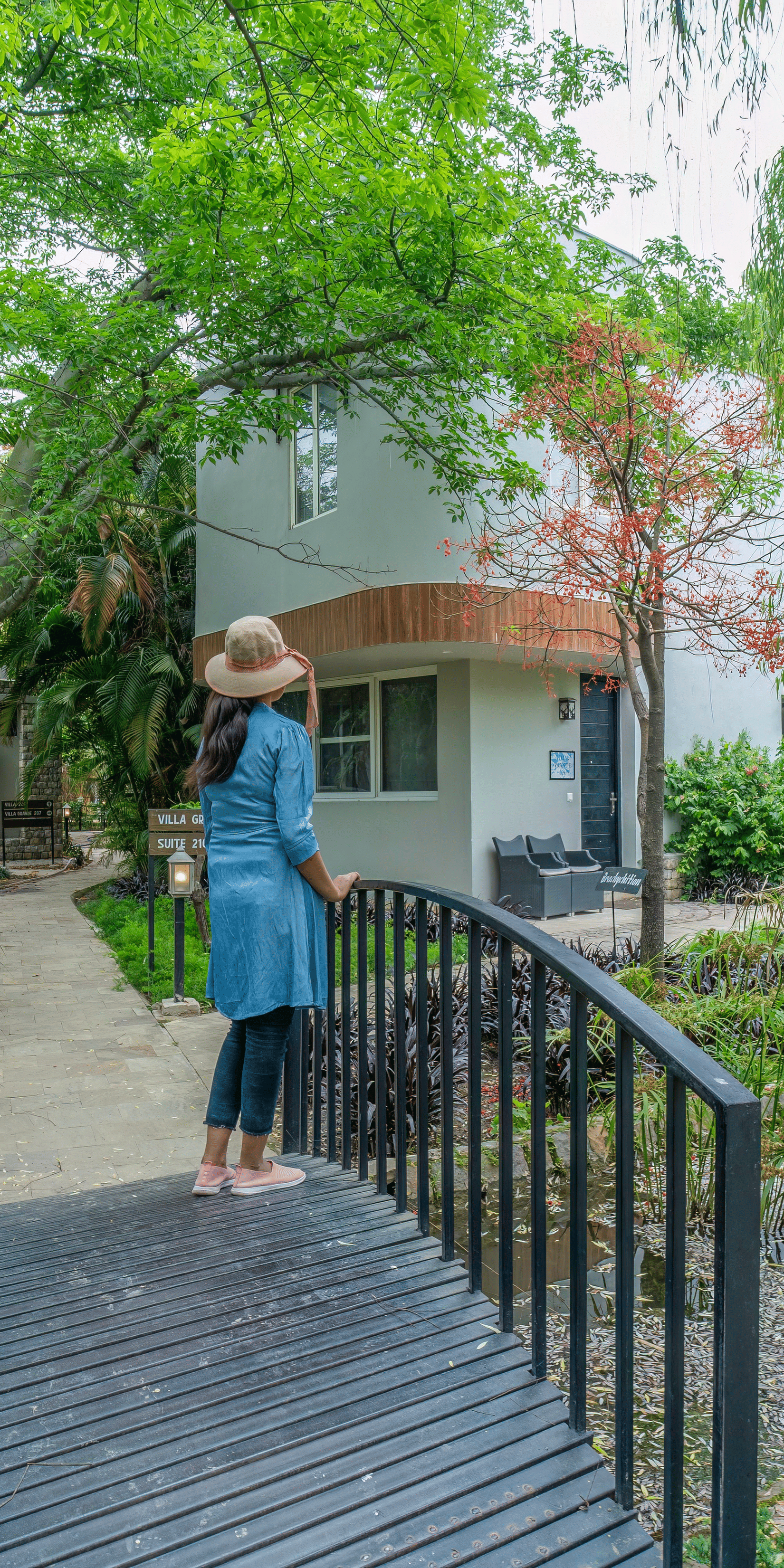 A bridge with a women standing on it which is over a small pond leading to a building surrounded by trees and greenery at The Golden Tusk, Jim Corbett