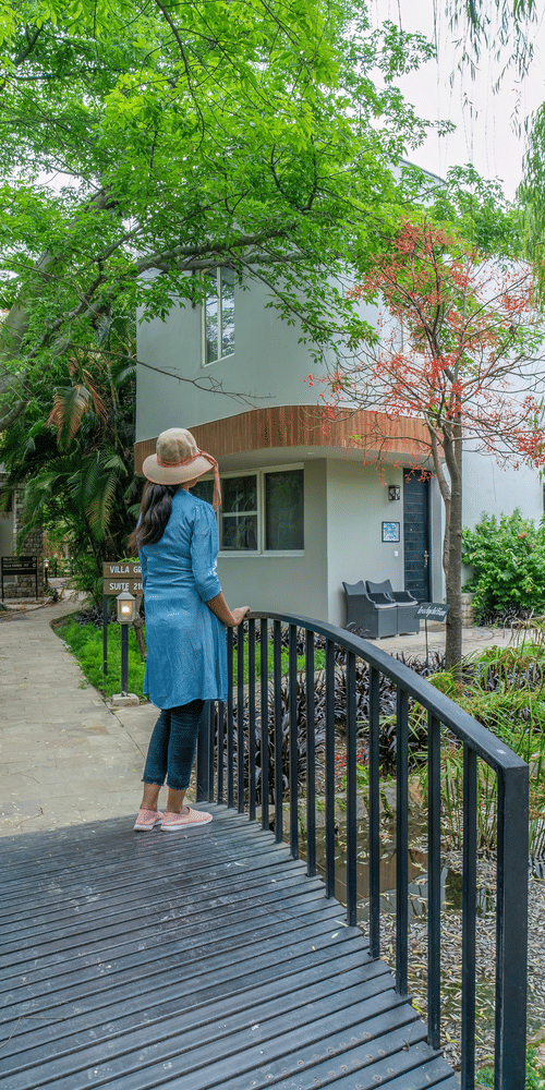 A bridge with a women standing on it which is over a small pond leading to a building surrounded by trees and greenery at The Golden Tusk, Jim Corbett