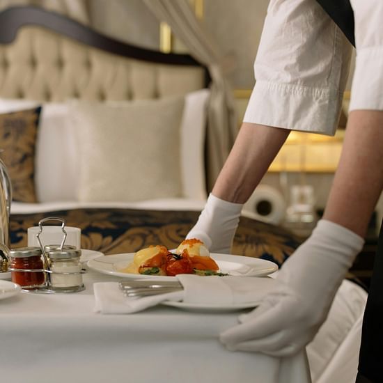 A hotel staff member in white gloves serving breakfast on a trolley next to an elegantly made bed.