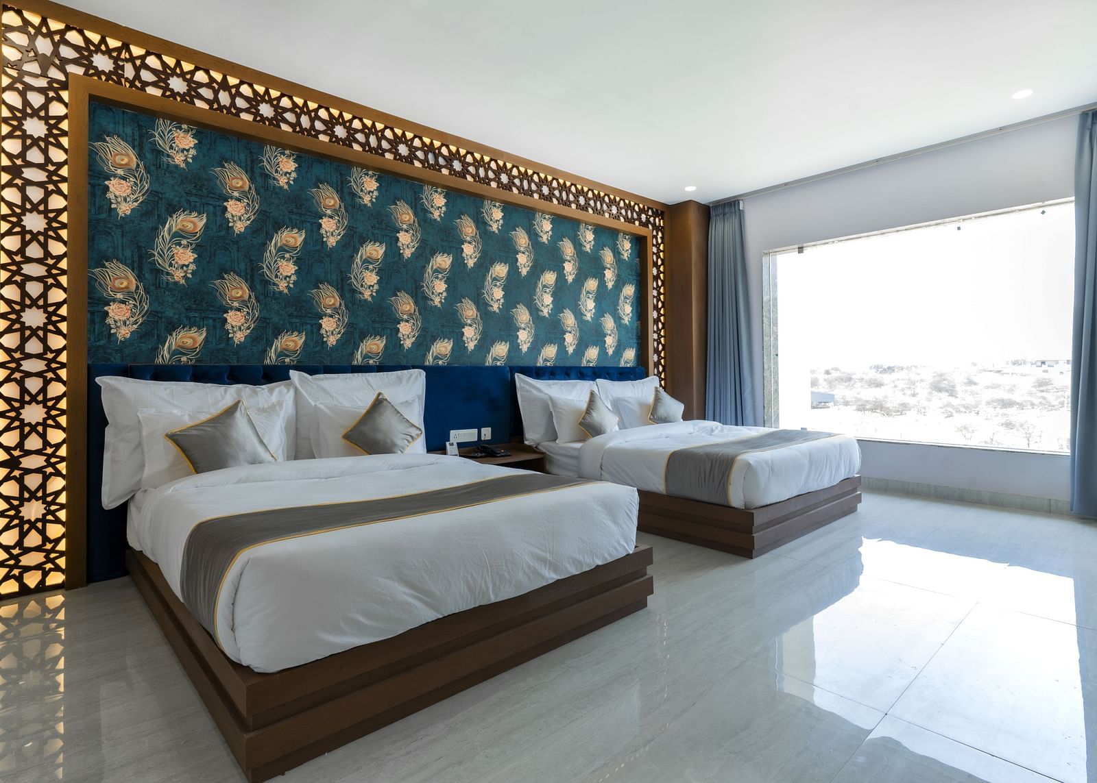 A bedroom at the Bamboo Saa Mulberry Resort featuring two twin beds with white linens and grey throws. A decorative wall panel with a blue and gold pattern is above the beds and a large windows is visible.