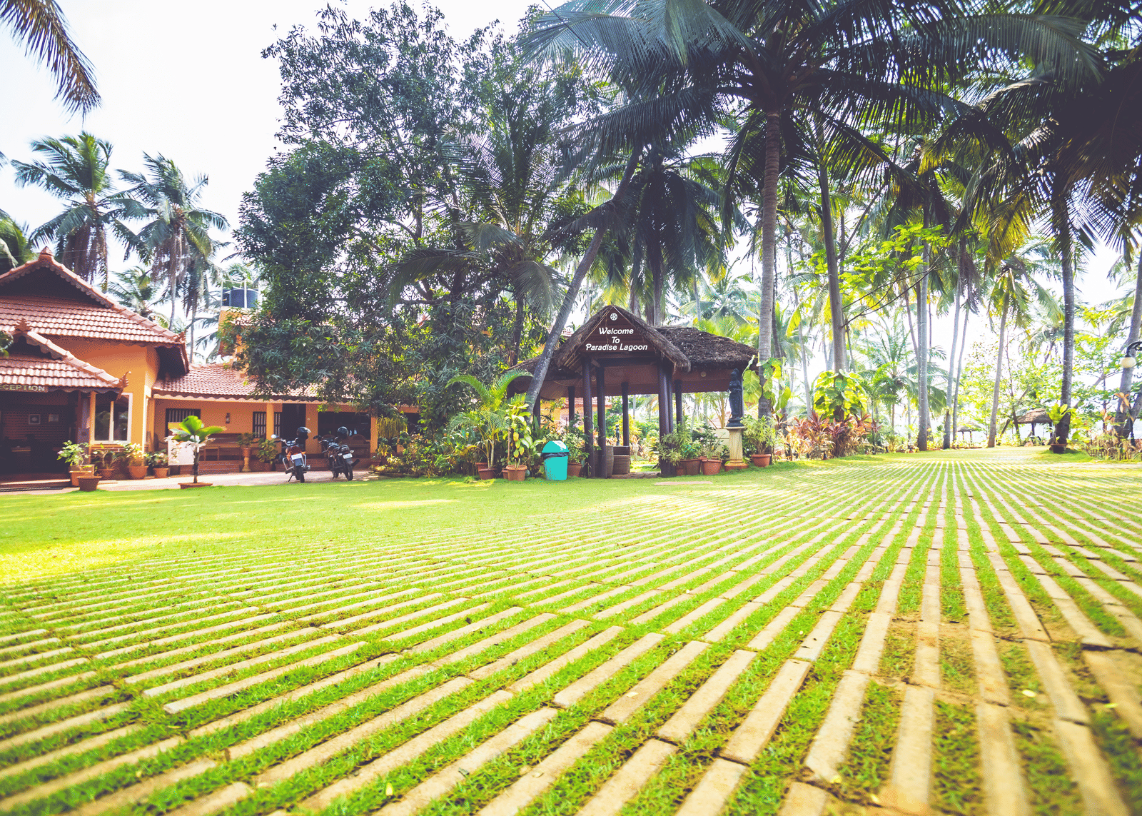 Lawn with palm trees, tiled pathways, and red-roofed building surrounded by greenery at Paradise Lagoon Resort, Udupi.