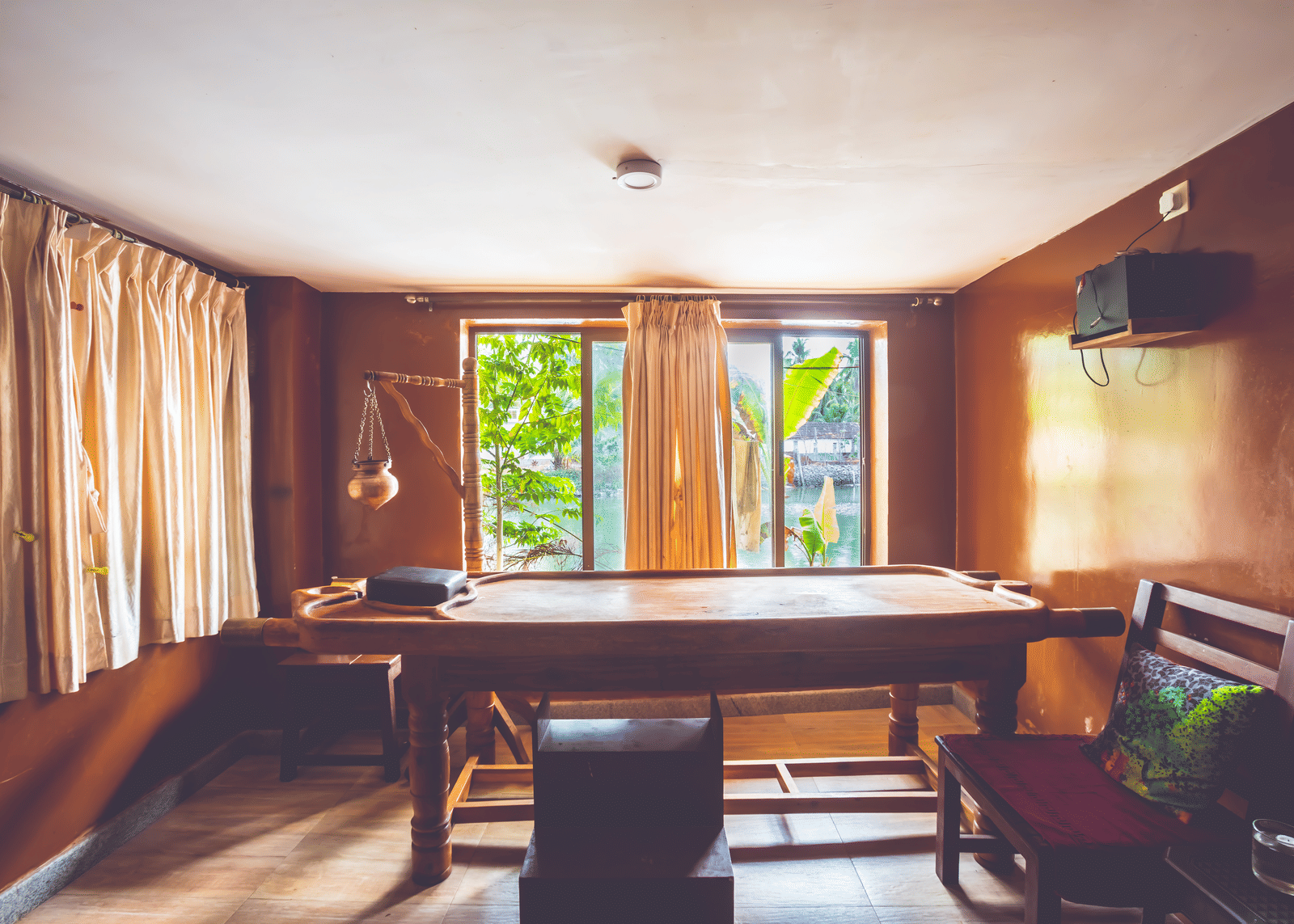 Room with wooden massage table, wooden chairs, desk, and curtained windows letting in natural light from both sides at Paradise Lagoon Resort, Udupi.