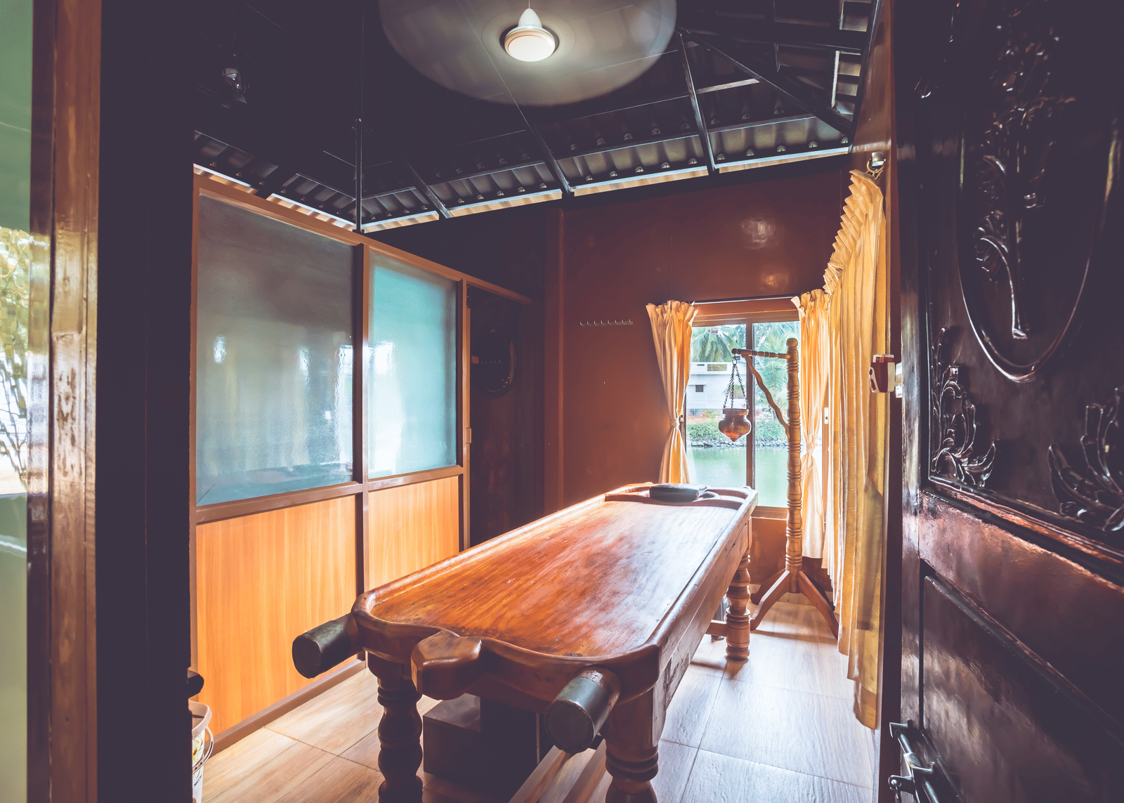 Room with a traditional massage table, ceiling light, and glass-panelled windows at Paradise Lagoon Resort, Udupi.