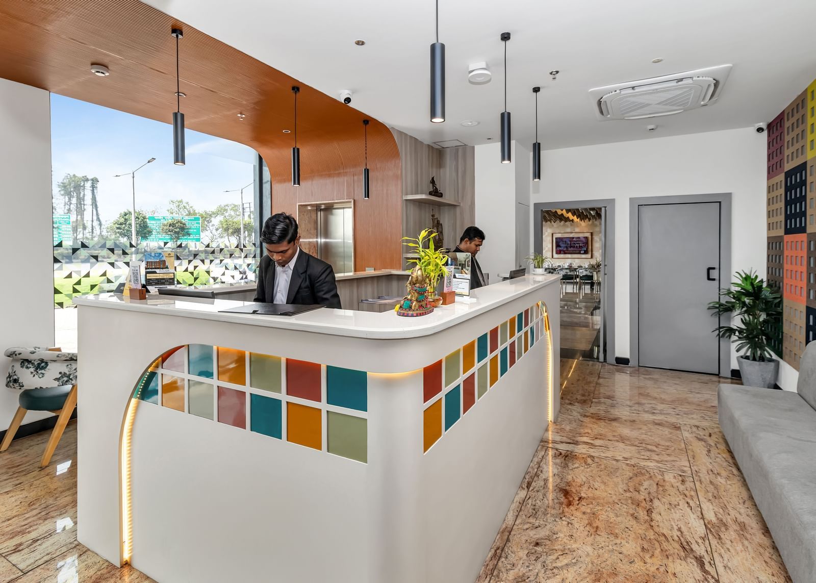 A brightly lit hotel reception area with staff behind a colourful tiled counter and a grey sofa in the waiting area | The Citi Residenci Hotel Banskopa