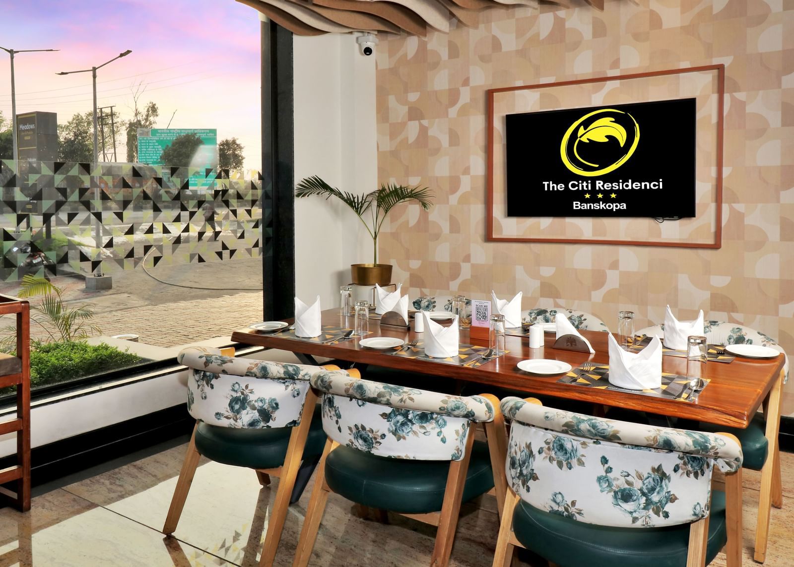 A restaurant dining area with a long wooden table set for guests, surrounded by chairs with floral upholstery, and a framed logo on the textured wall | The Citi Residenci Hotel Banskopa