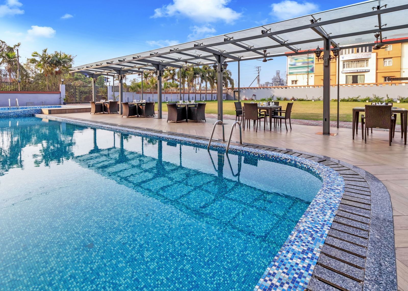 A sparkling blue outdoor swimming pool with mosaic tiling along the edge, surrounded by a paved area and covered dining tables under a clear sky with scattered clouds  | The Citi Residenci Hotel Banskopa