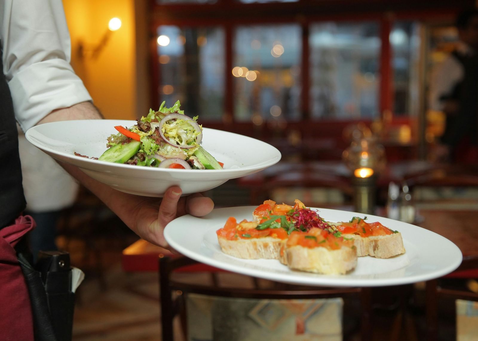 A waiter in a white shirt and black vest carefully carries a plate of salad and a plate of bruschetta in a warmly lit restaurant setting.