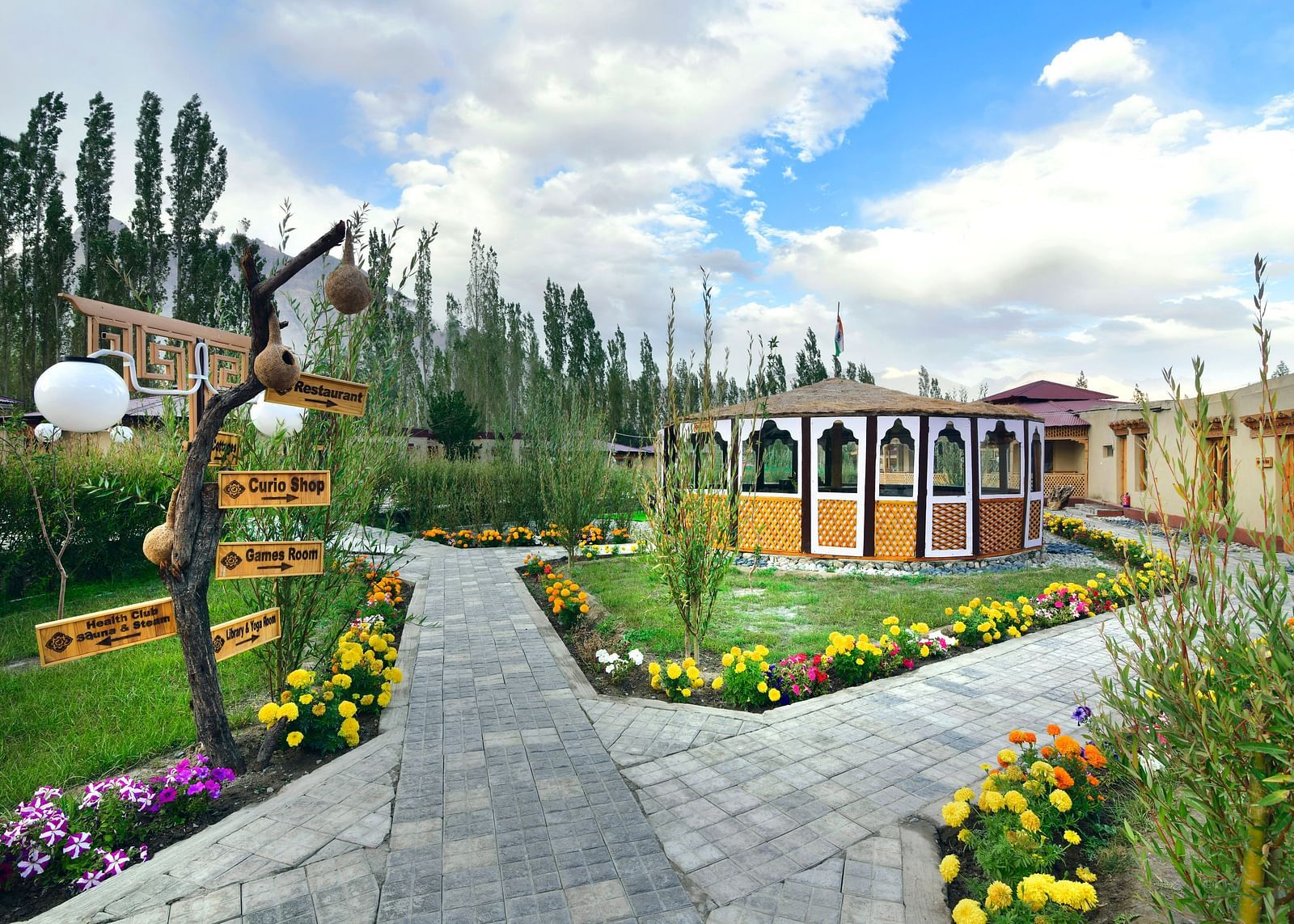 A pathway leading to White Sands Resort, Ladakh lined with flowers and greenery, with decorative signage and accommodation buildings visible against a backdrop of trees and a cloudy sky.