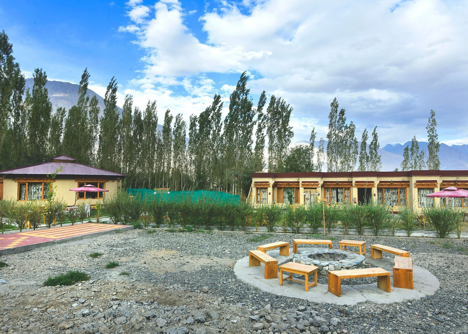 An overview of White Sands Resort, Ladakh featuring a communal seating area with wooden benches and a fire pit surrounded by gravel, with cottages and trees in the background under a cloudy sky.