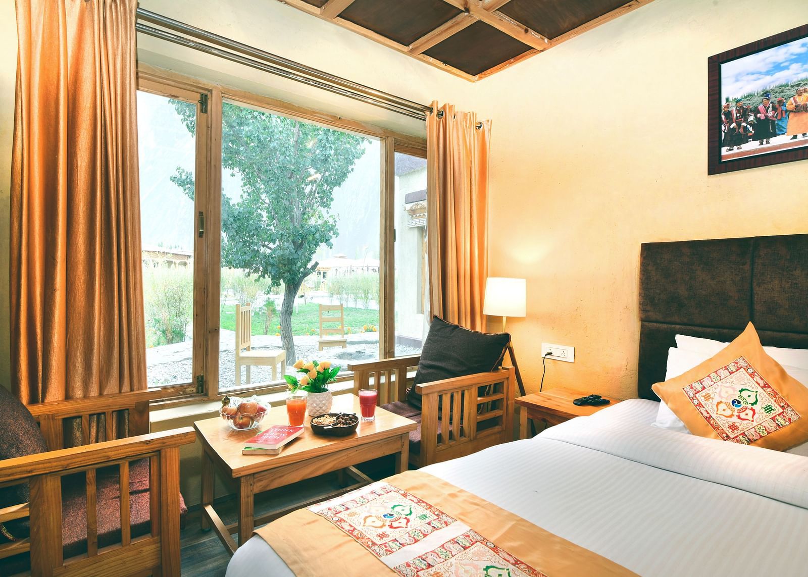 Standard Cottage with a king size bed, a window with orange curtains, a small table and chairs, and a painting on the wall at White Sands Resort, Ladakh.