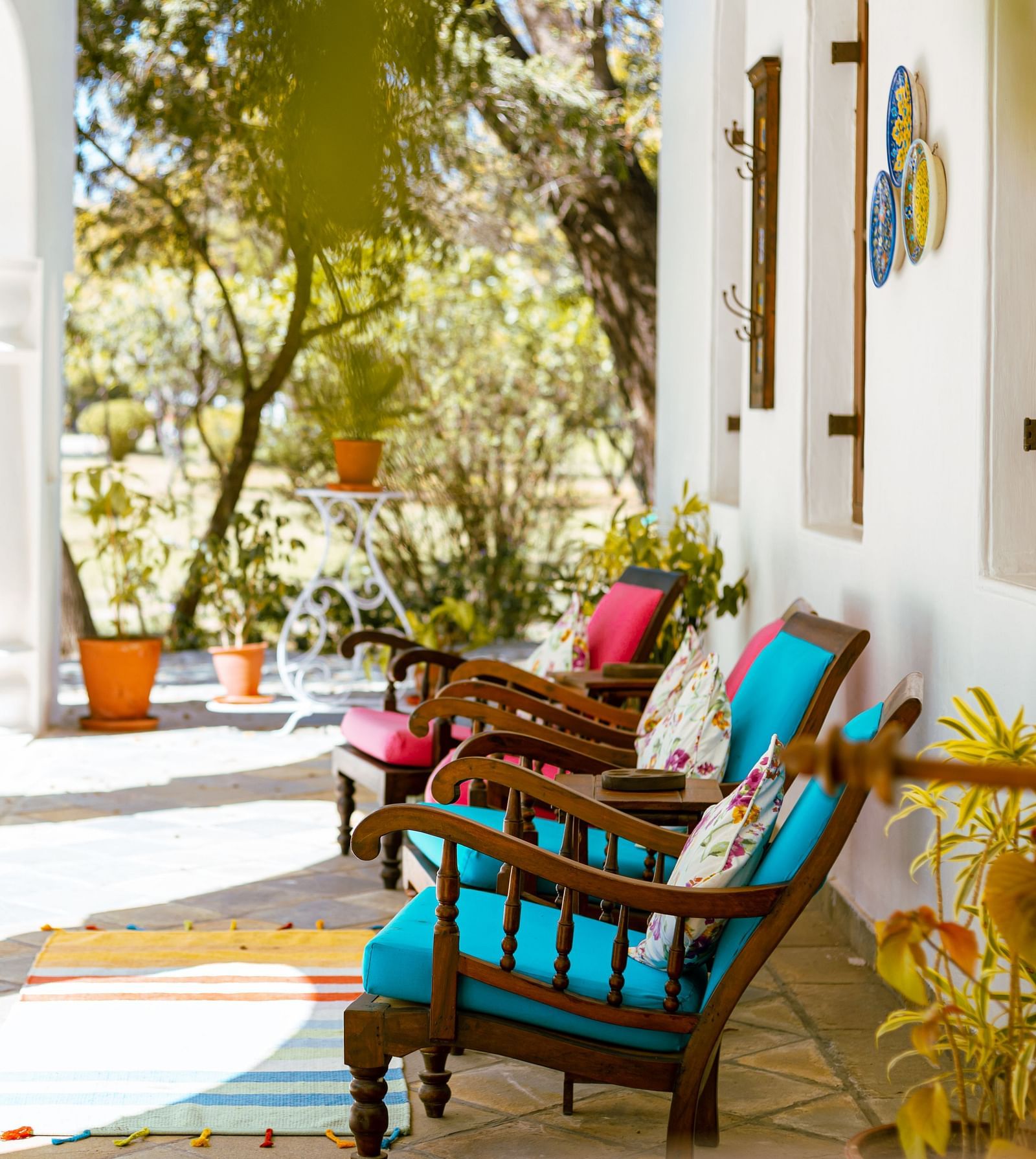 A bright, outdoor veranda with white columns and arches features colorful chairs with cushions, overlooking a garden with trees- Shahpura Bagh.