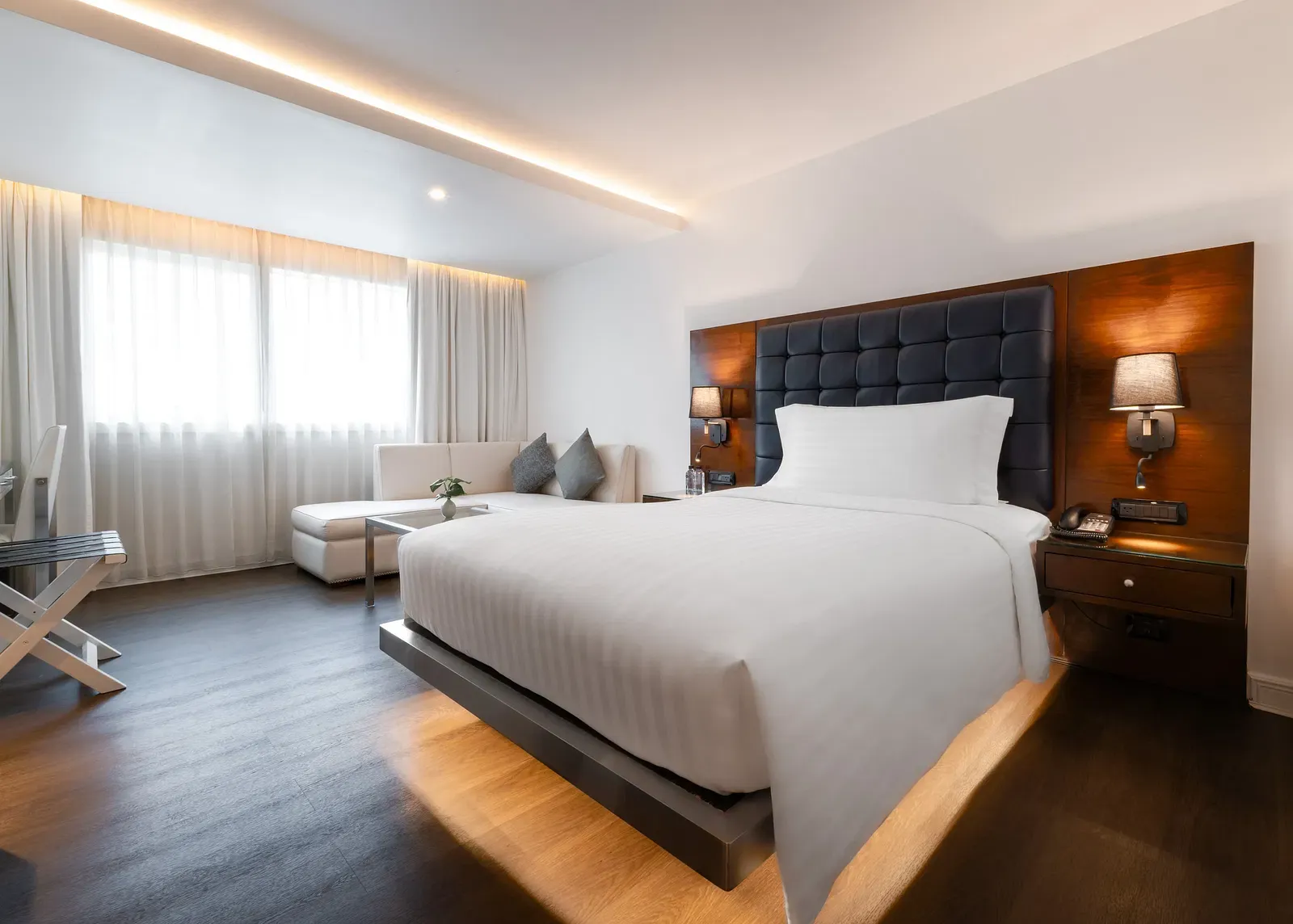 Modern hotel bedroom with tufted navy headboard, platform bed, white seating area, walnut accents, and floor-to-ceiling curtains at Night Hotel Bangkok.
