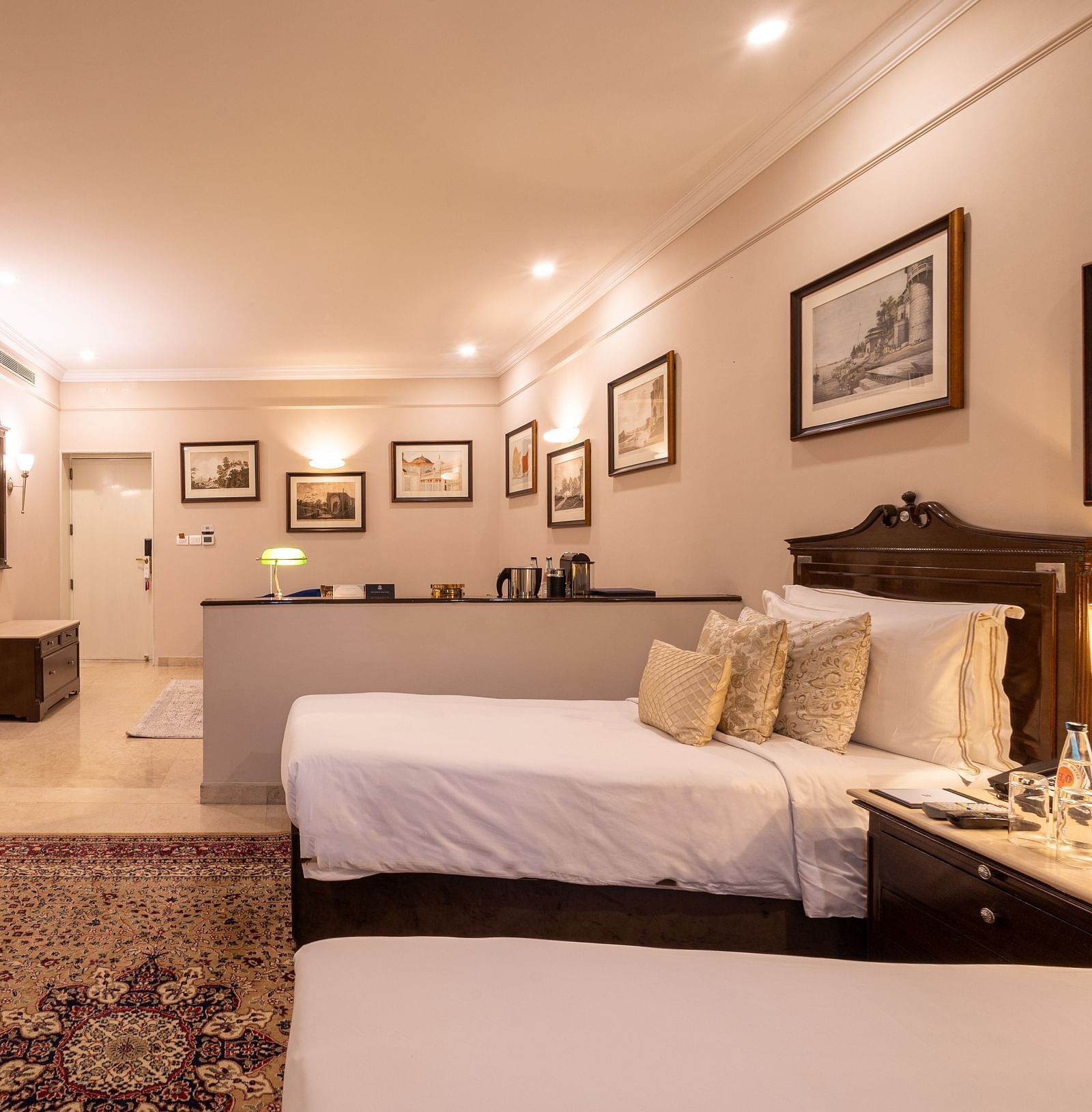 Side view of the Grand Luxury Twin Bed Room at The Claridges New Delhi, showcasing a spacious layout with framed photos on the wall and an elegant lighted ceiling
