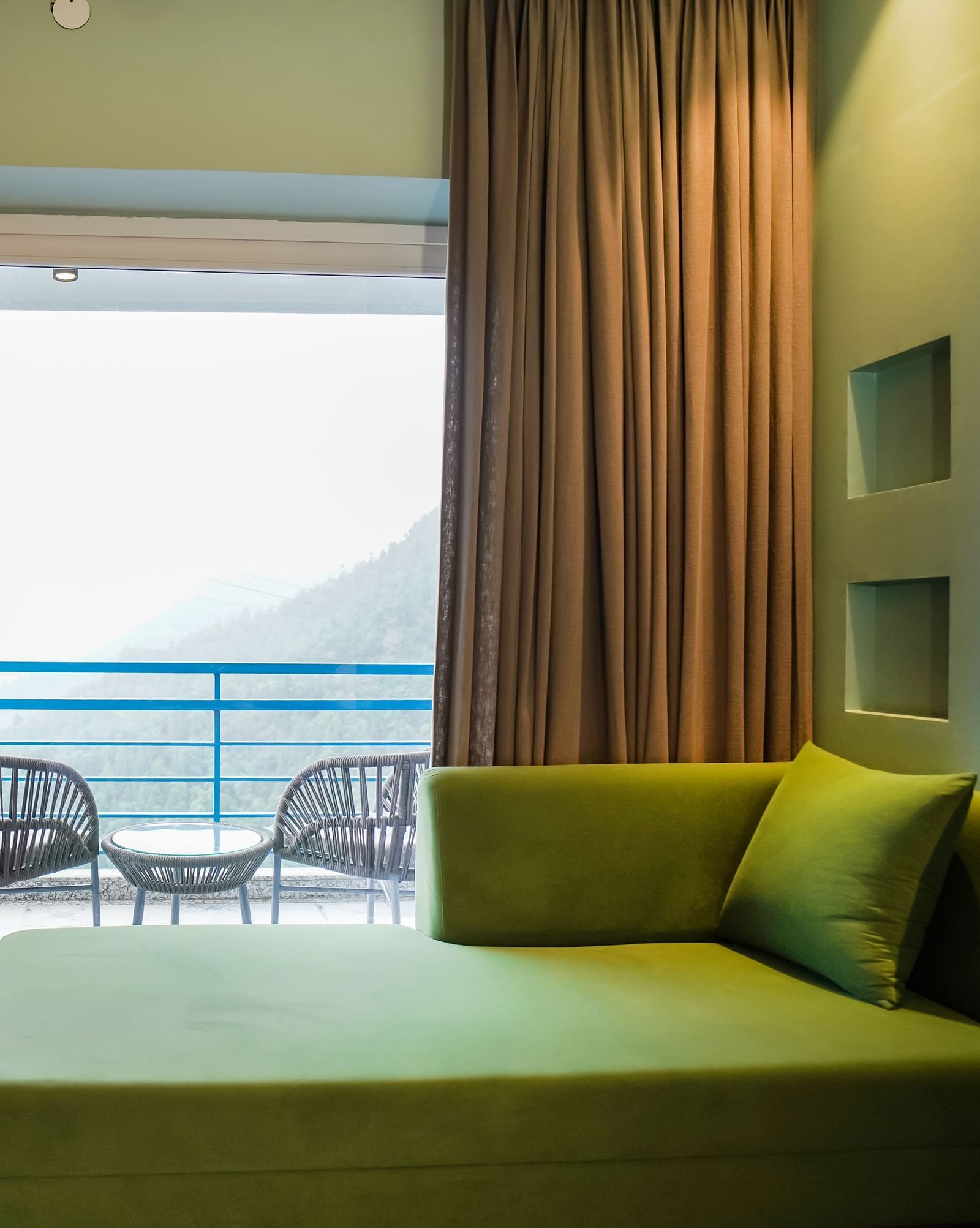 A modern hotel room with a green sofa and a large sliding glass door opening to a balcony with outdoor seating and a view of the hills | Deluxe Room with Valley View | Sukama Resort & Spa, Nainital