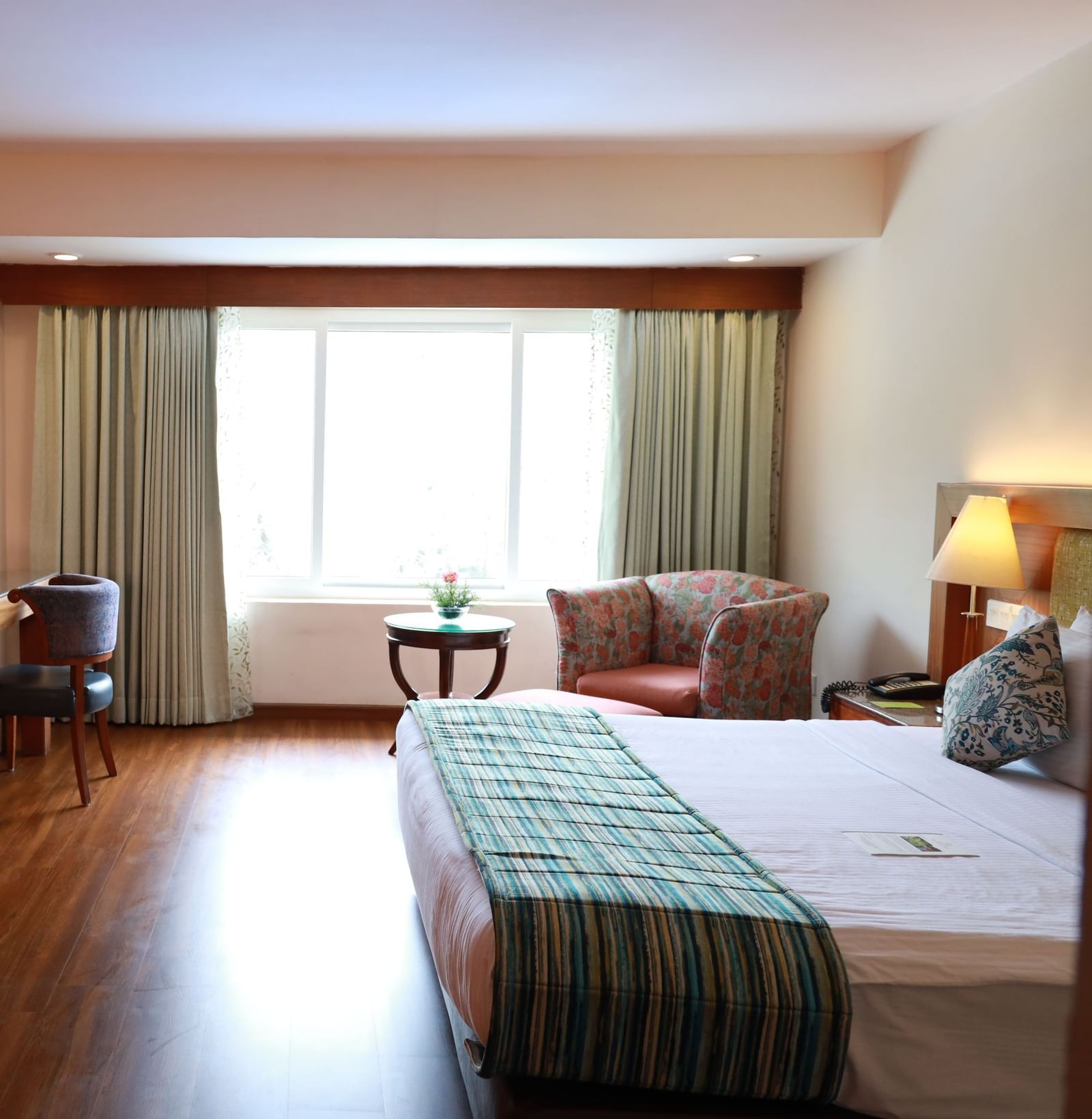 A well-lit room at Accord Highland Ooty featuring a desk, chair, and a plush bed with a green throw.