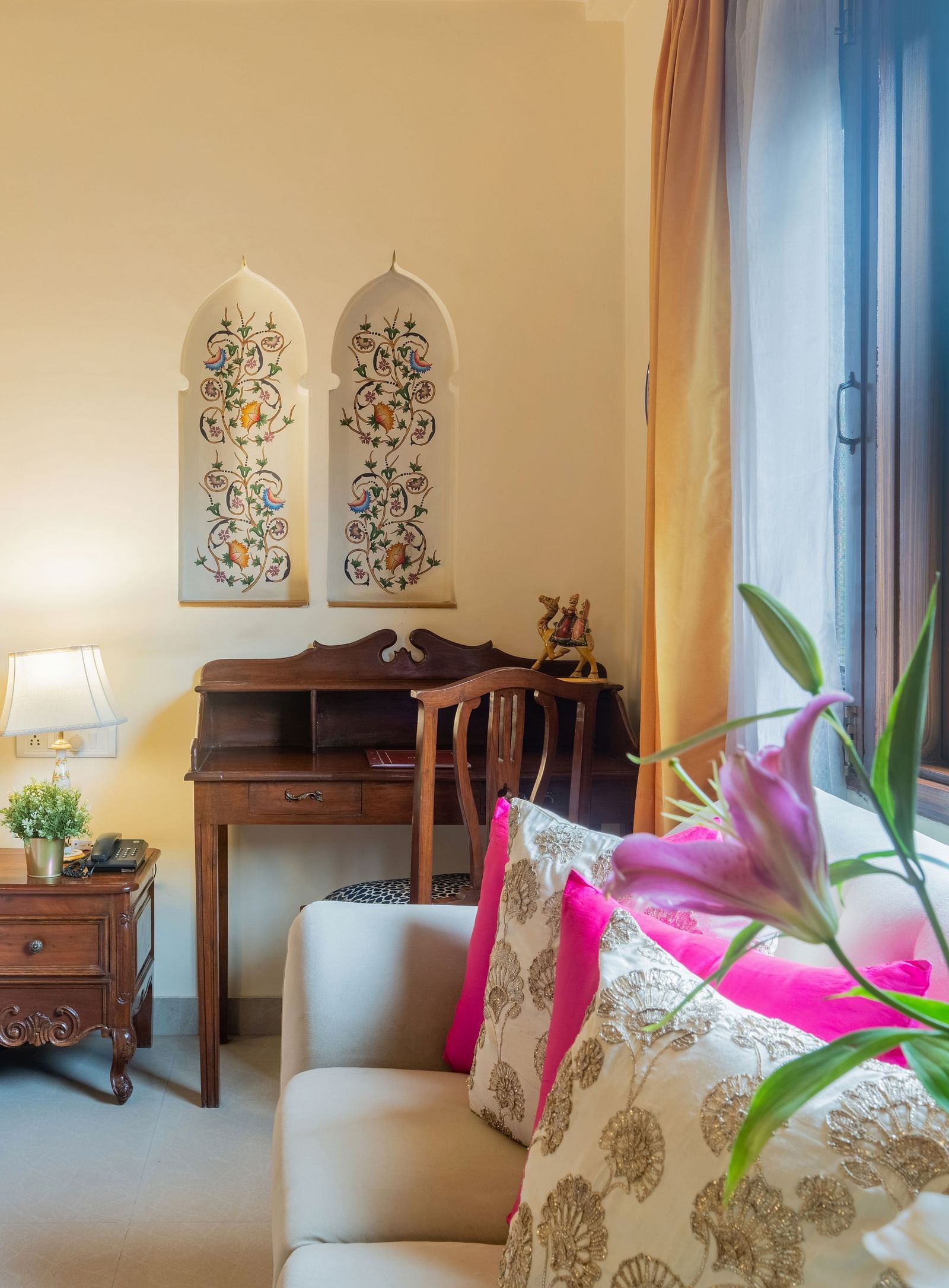 A lovely room at Dera Rawatsar, Jaipur, features a four-poster bed, a comfortable sofa, a writing desk, and decorative floral accents on the wall.