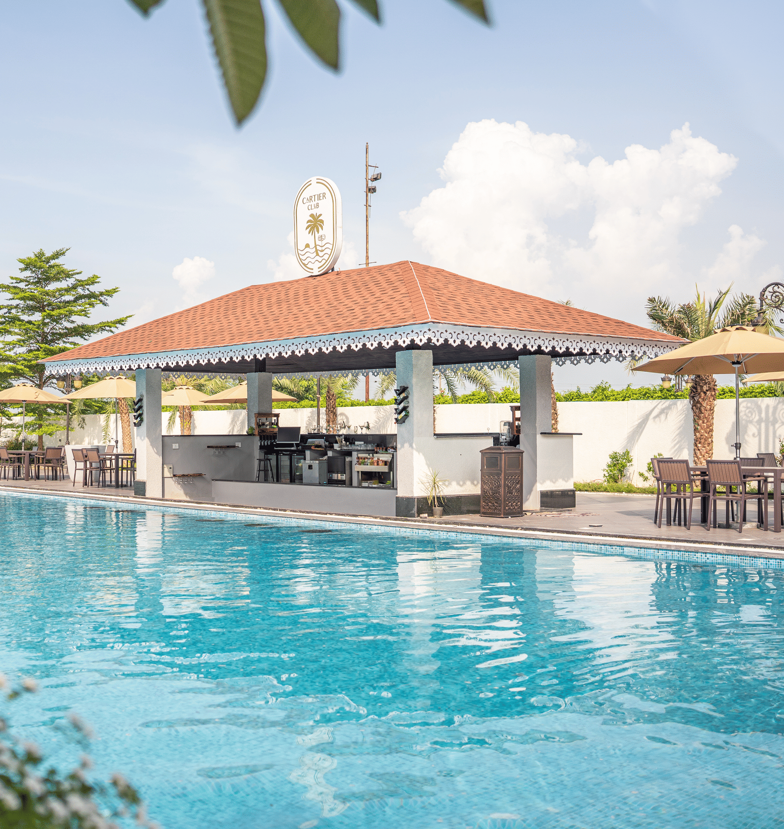 Sunny daytime view of the swimming pool with a red-roofed bar or pavilion and lush greenery in the foreground at Hotel Hukam's Lalit Mahal.