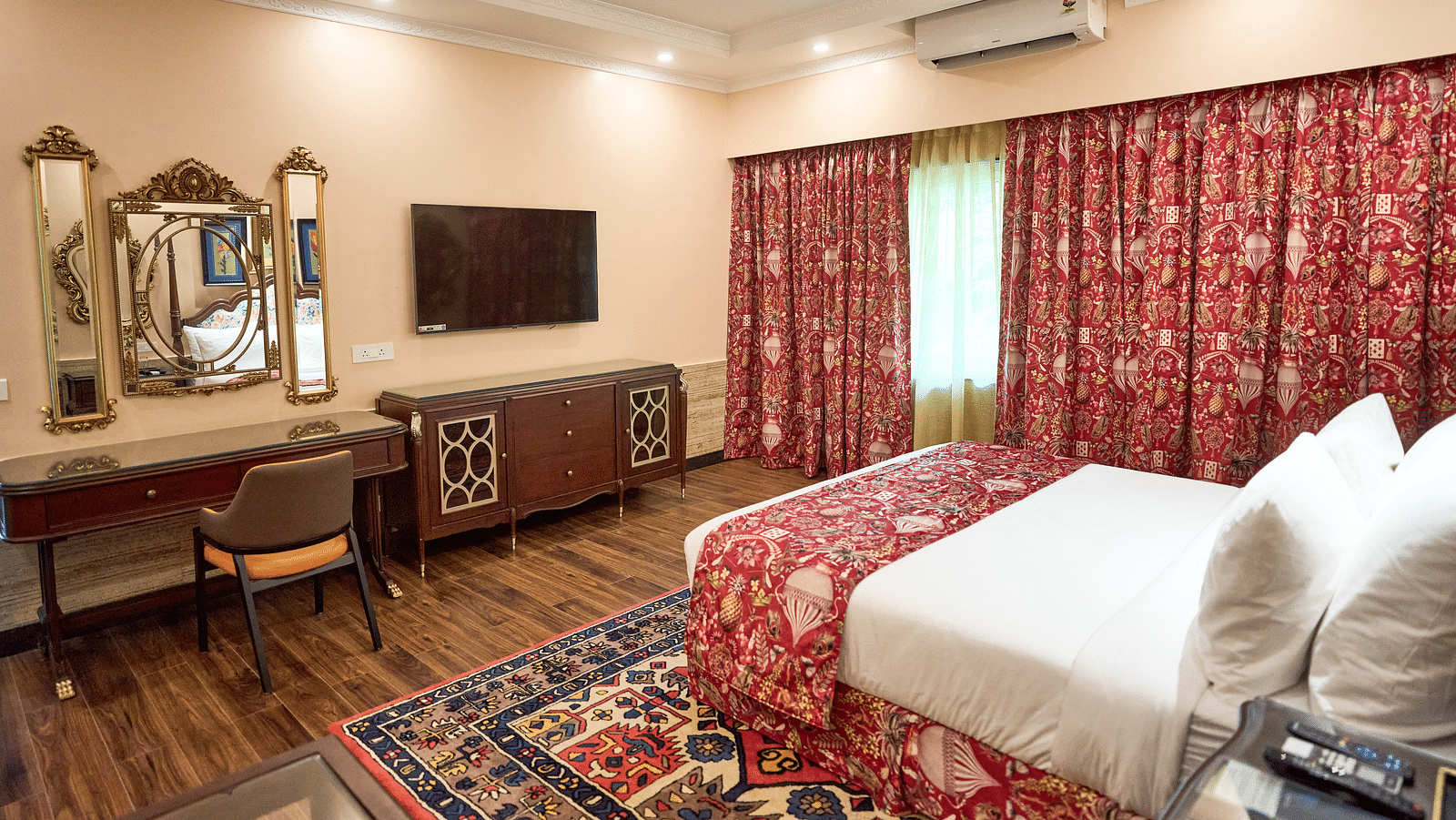 A spacious room featuring a dressing table with an adjacent mirror, a wall-mounted TV above a wooden table, captured from a bedside perspective at Mayfair Manor, Alibag