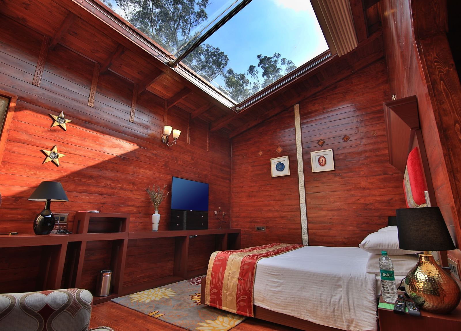 A wide angle image of a room in Ooty featuring a bed, a television and a glass roof flooding the space with sun light - Accord Highland Ooty