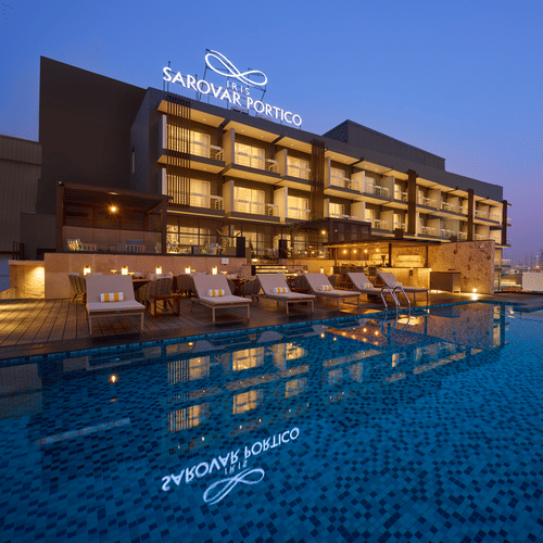 A modern hotel building is illuminated at dusk, featuring a rooftop swimming pool reflecting the hotel's signage and nearby lounge chairs. - Iris Sarovar Portico, Sector 85, Gurugram