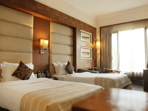 Deluxe Rooms at The Bristol Hotel Gurgaon, Rooms in Gurgaon, featuring twin beds, spreads, pillows, tables, and a window coveed with curtains.