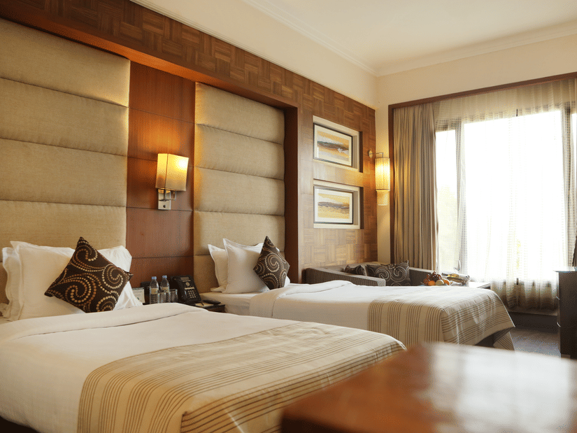 Deluxe Rooms at The Bristol Hotel Gurgaon, Rooms in Gurgaon, featuring twin beds, spreads, pillows, tables, and a window coveed with curtains.