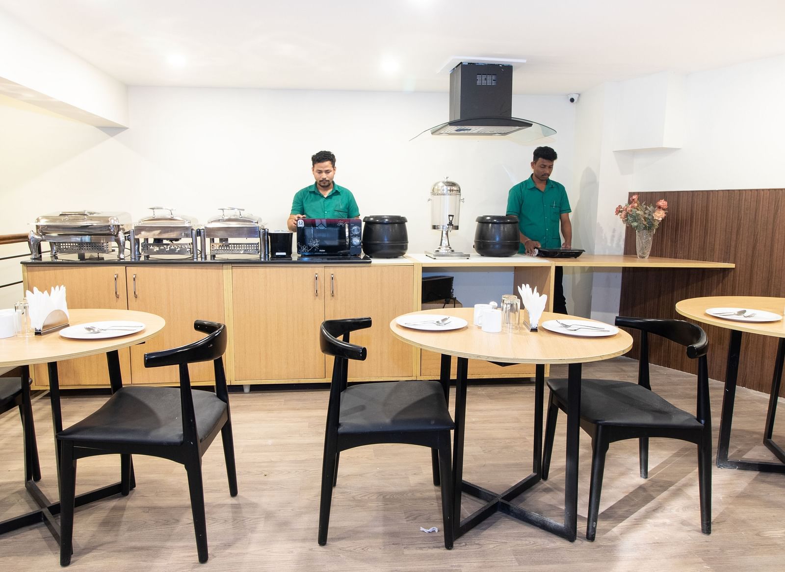 A breakfast buffet area with staff behind the counter, food warmers, and tables with black chairs - Sanctum Suites Richmond Road, Bangalore