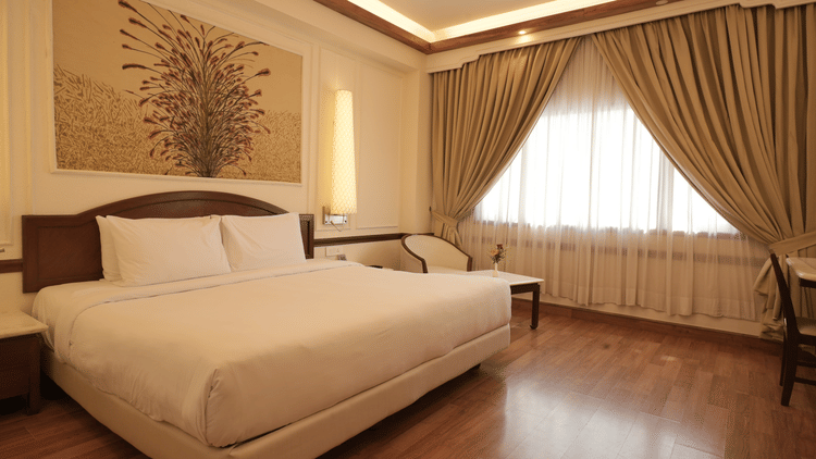Polished hardwood floors leading towards a bed and large window with draped curtains in the Superior King room at Clarks Amer, Jaipur.