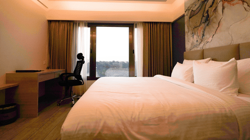 A bright hotel room featuring a double bed, a desk and chair, and a large window or balcony door | Pride Elite Chhatrapati Sambhajinagar