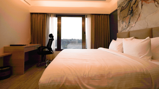 A bright hotel room featuring a double bed, a desk and chair, and a large window or balcony door | Pride Elite Chhatrapati Sambhajinagar