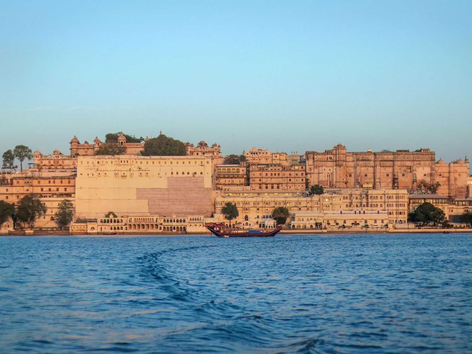 a far out view of city palace in udaipur with a waterbody in front of it.