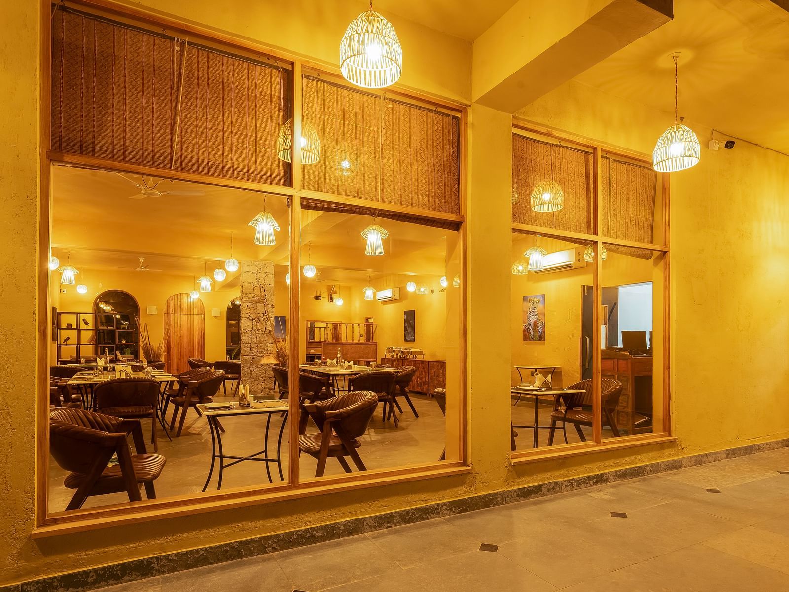 An image of L cave restaurant where walls are made of glass and it has a boho theme featuring several sets of dining tables and chairs - Kurja Jawai