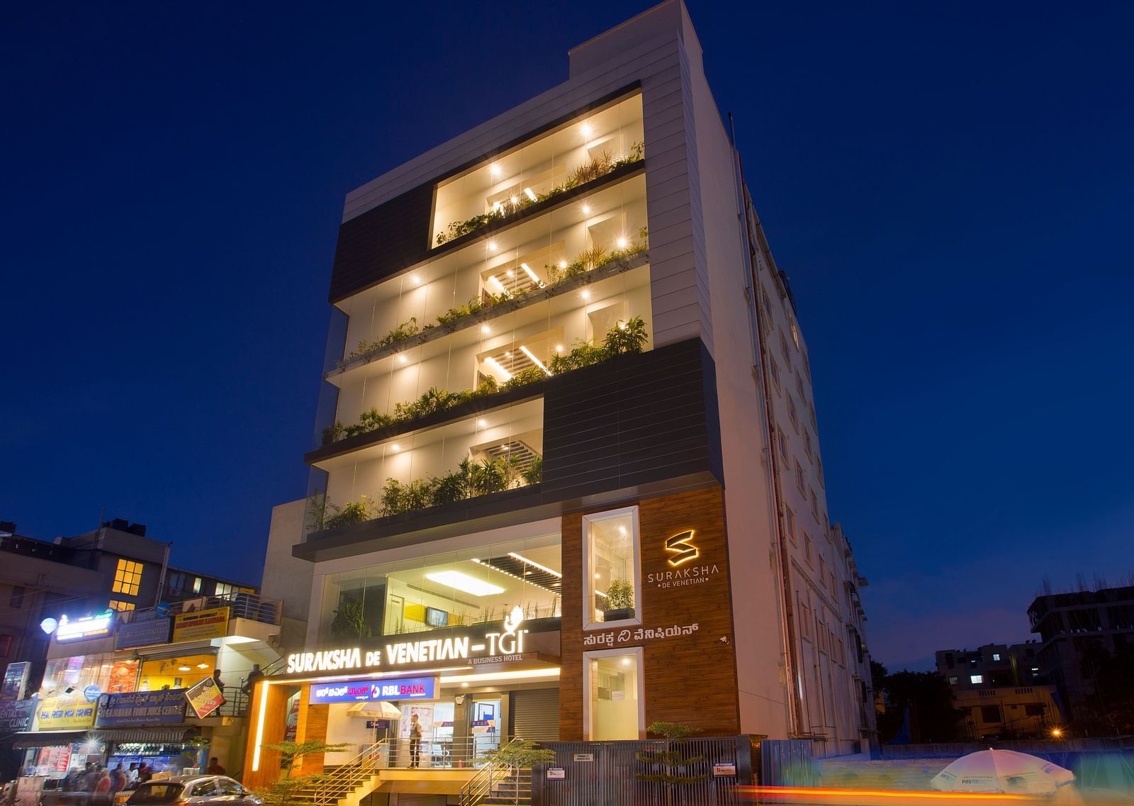 An exterior view of a modern multi-story building illuminated at dusk with signage and street activity - De Venetian by TGI, Brookefield