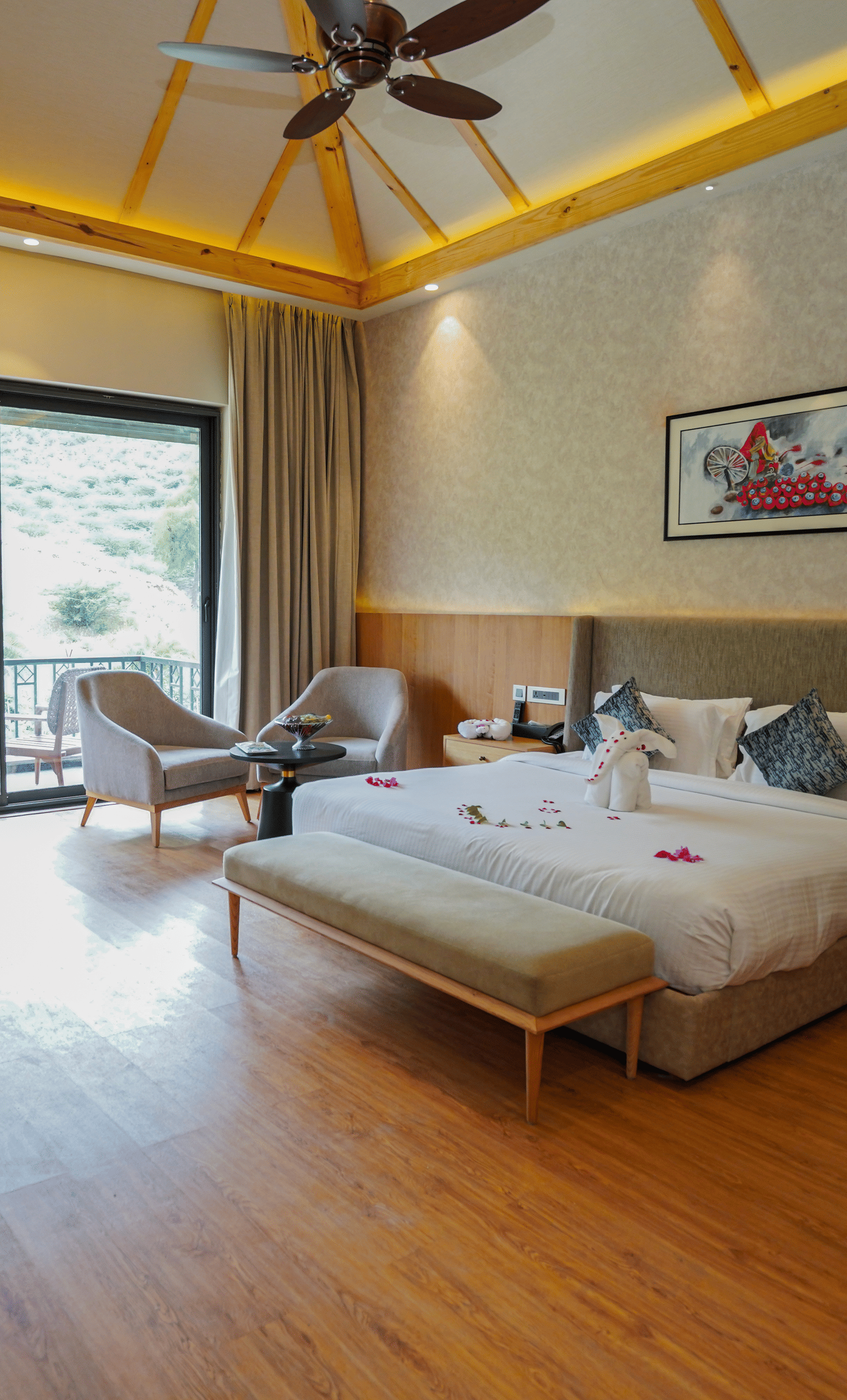 A modern hotel room with wooden floors and a large bed covered in rose petals, a desk, and a large window overlooking a balcony.
