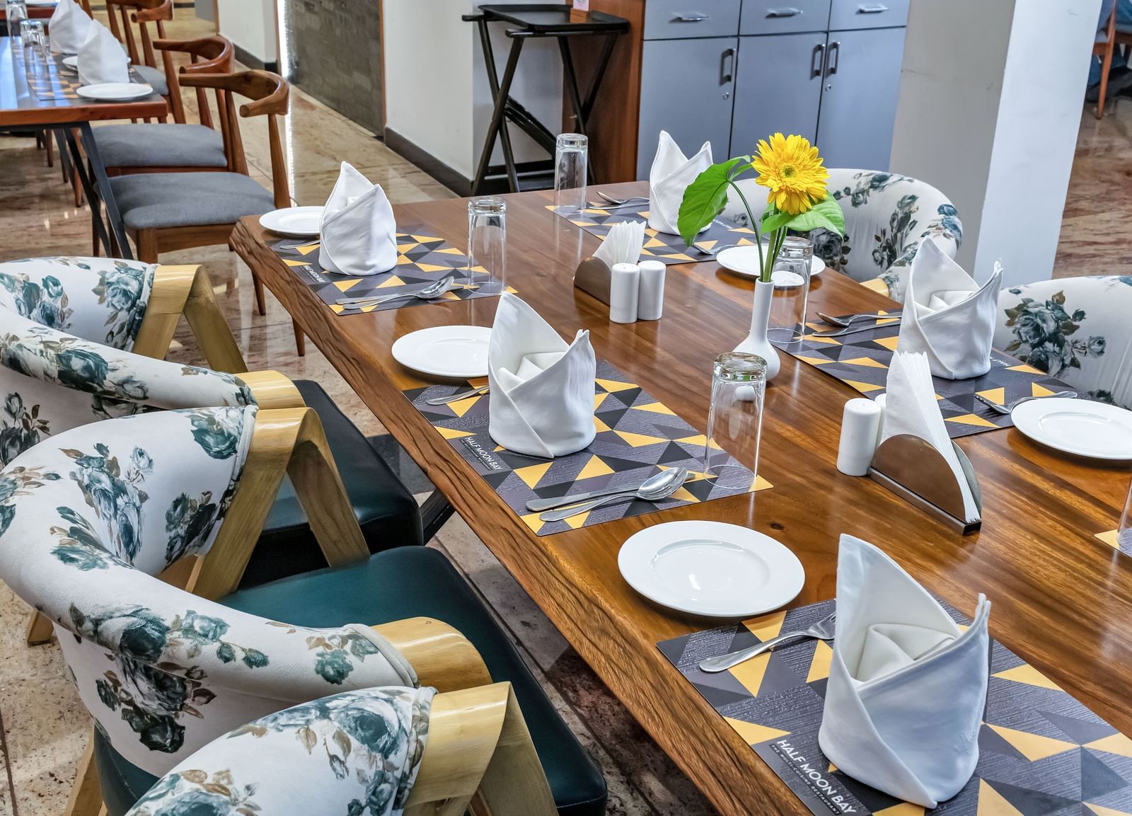 A long wooden dining table set with plates, folded napkins, glasses, and a yellow flower centerpiece, surrounded by chairs with floral patterned upholstery | The Citi Residenci Hotel Banskopa
