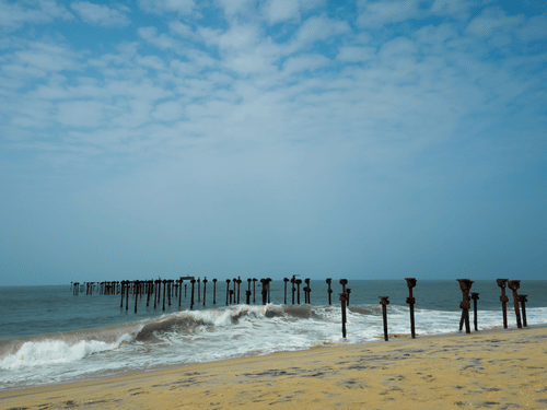 Weathered wooden sea pier posts stand in breaking surf along a sandy shoreline beneath a bright, cloudy sky.