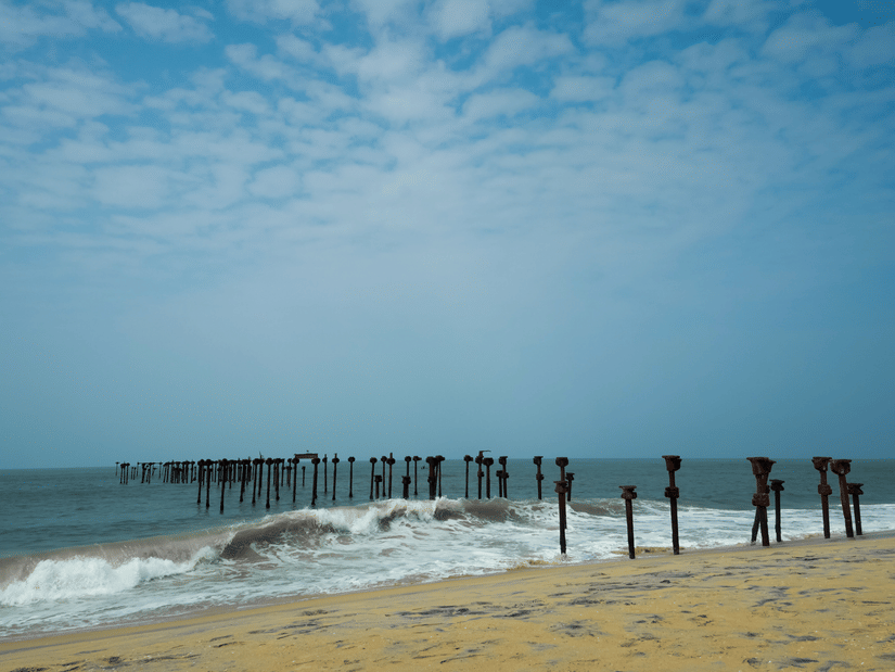 Weathered wooden sea pier posts stand in breaking surf along a sandy shoreline beneath a bright, cloudy sky.