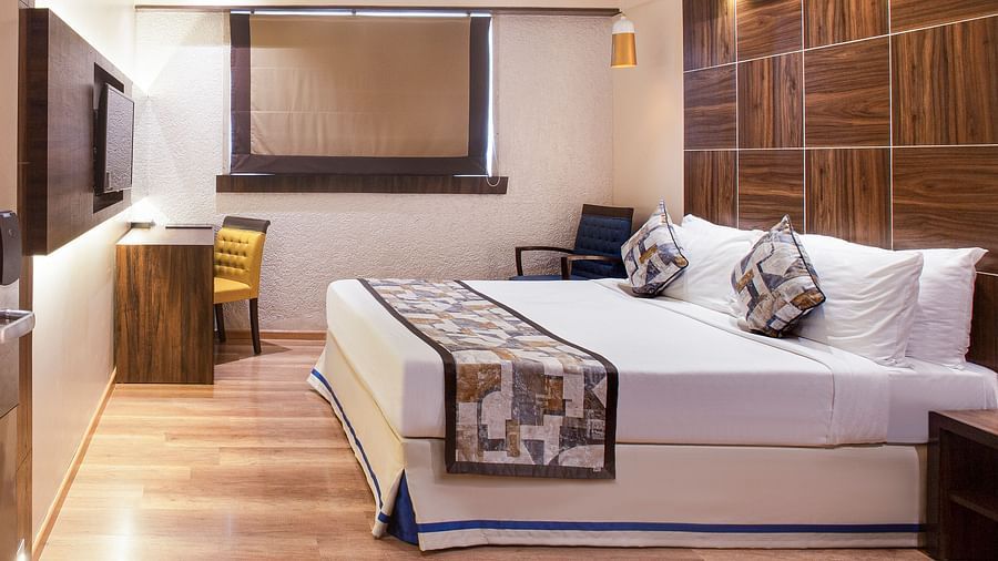 A side angle view of the Deluxe room at Hotel Bawa Regency, Dadar, featuring a comfy bed, a work desk, and a window.