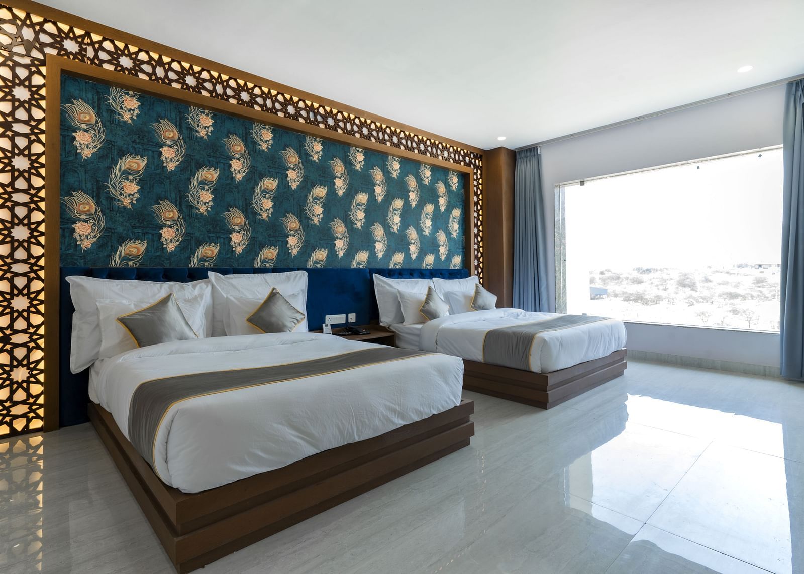 A bedroom at the Bamboo Saa Mulberry Resort featuring two twin beds with white linens and grey throws. A decorative wall panel with a blue and gold pattern is above the beds and a large windows is visible.